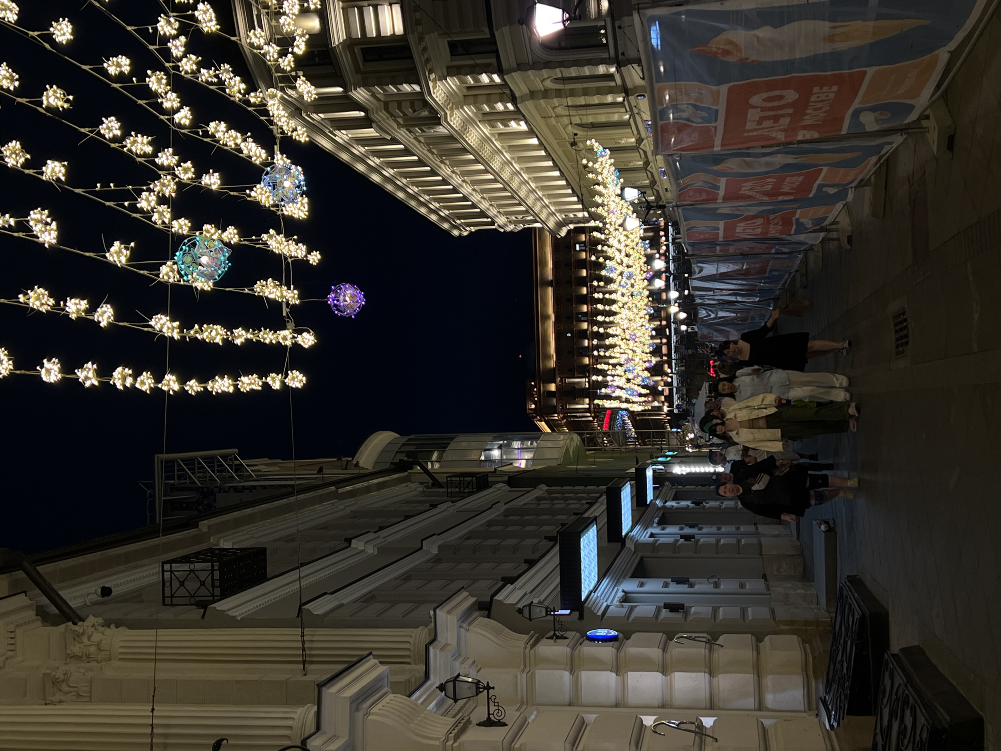 The image shows a street scene in Moscow, Russia, at night. The street is illuminated by strings of decorative lights with spherical accents, creating a festive atmosphere. Buildings line both sides of the street, displaying ornate architectural details. A group of people walks along the sidewalk on the right side of the frame. Banners are hung above the sidewalk.