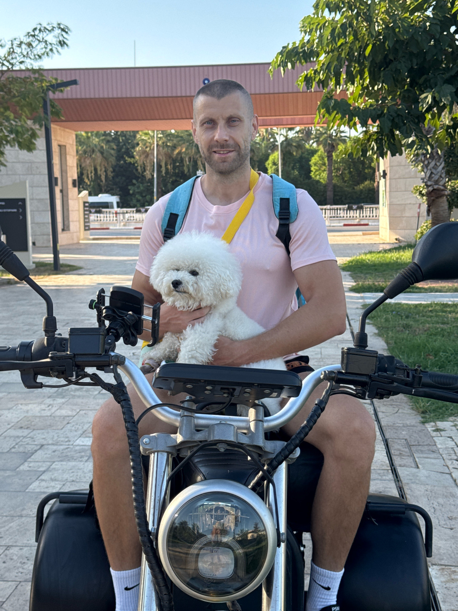 A man with short hair and a beard sits on an electric scooter with a white Bichon Frise dog in his arms. He is wearing a pink t-shirt, white socks with Nike logos, and dark shorts. The dog is fluffy and white with a black nose. The man is wearing a blue backpack with a yellow strap. He is looking at the camera with a slight smile. The scooter has a silver handlebar and a black headlight. The background shows a paved walkway, trees, and a building with a reddish-brown roof. The scene appears to be in an outdoor area, possibly a resort or a park, in Antalya, Turkey, given the architecture and vegetation typical of the region.