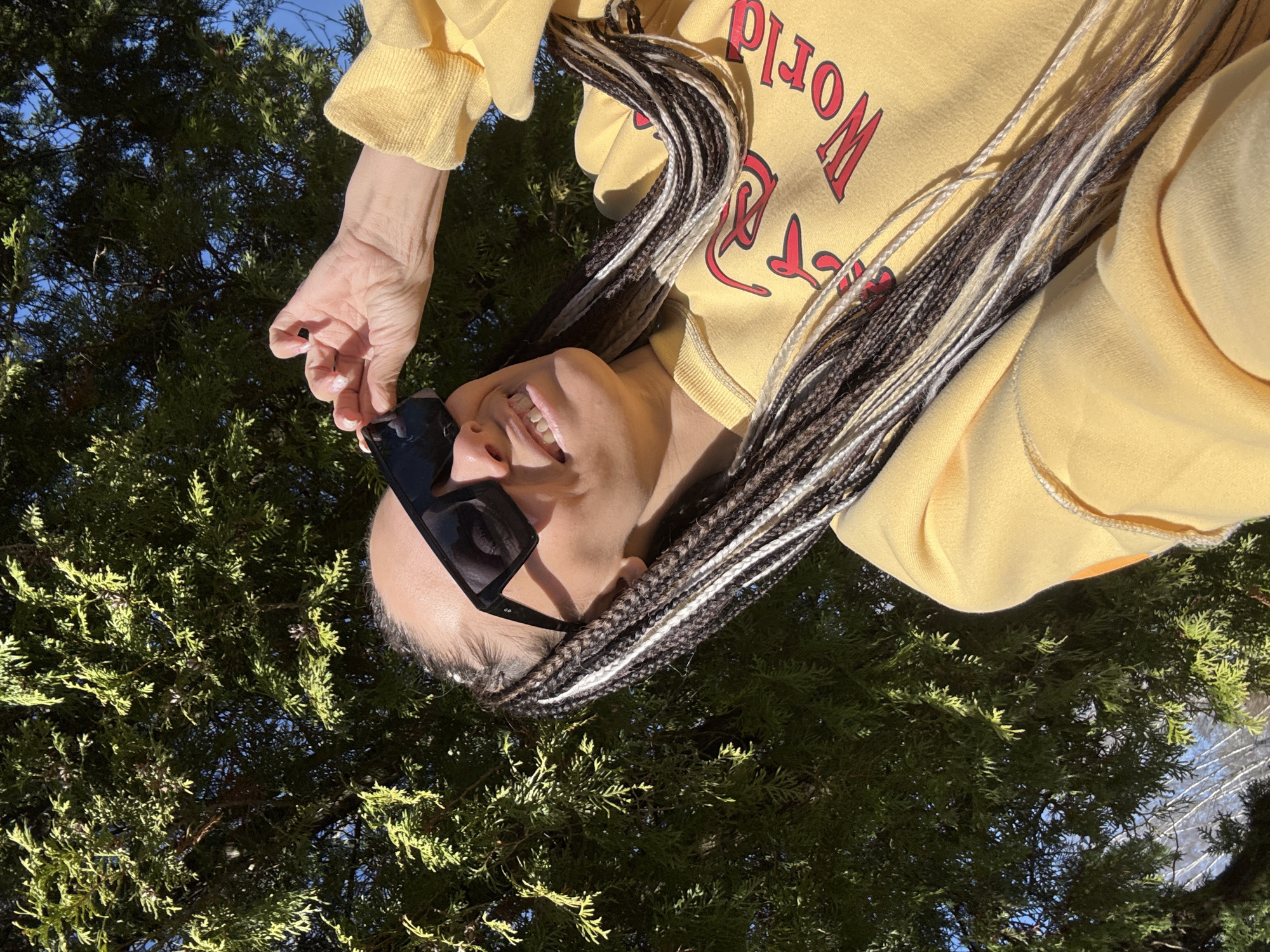 A person is upside down in an outdoor setting. The person is wearing a yellow sweatshirt with red lettering, braids of mixed brown and white, and sunglasses. The person is smiling and holding onto their sunglasses with their left hand. The background consists of green foliage and a glimpse of a blue sky. The location is indicated as the city of Rusanovo, Russia, but no city-specific features are distinctly visible in the image.