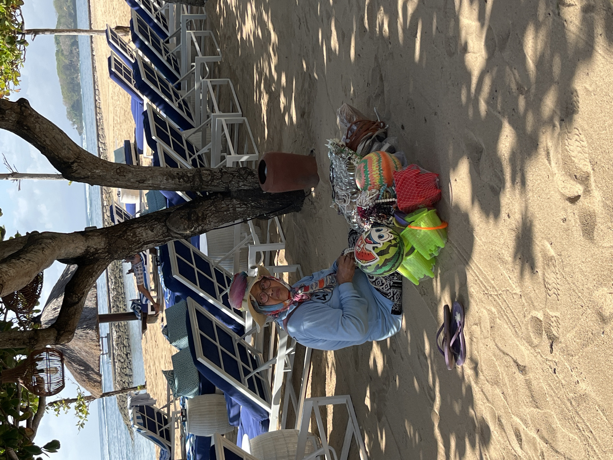 In Kuta Selatan, Indonesia, a vendor sits on a sandy beach under the shade of trees, surrounded by colorful items for sale. The vendor, wearing a light blue long-sleeved shirt, a patterned headscarf, and a straw hat, is seated on the sand with a pile of goods in front of them. The merchandise includes an assortment of small toys and accessories, such as buckets, nets, and beaded necklaces, displayed in a somewhat organized manner.
Behind the vendor, rows of empty blue lounge chairs with white frames are arranged on the sand, facing the ocean. A thatched umbrella provides additional shade in the background, and a stone barrier separates the beach from the water. The sea is calm with gentle waves, and a distant island or coastline is visible on the horizon. The overall atmosphere is one of a peaceful beach resort setting, with the vendor actively participating in the commercial activity of the location. A pair of purple flip-flops rests on the sand to the left of the vendor.