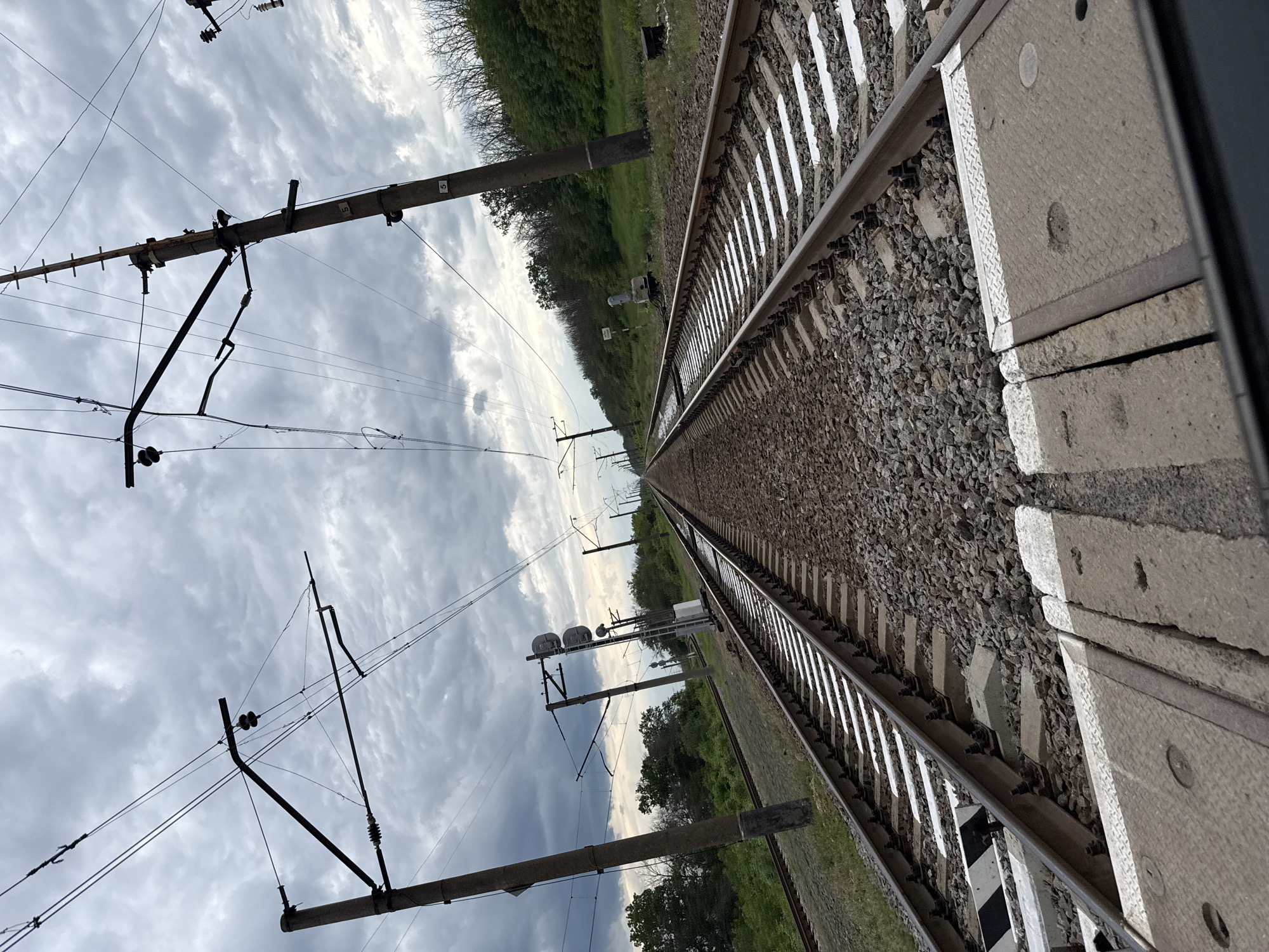 The image displays a straight section of a railway line extending into the distance under an overcast sky with scattered clouds. The perspective is from alongside the tracks, looking forward along their path. At least two parallel tracks are visible, supported by concrete sleepers and surrounded by gravel ballast.
Overhead catenary system poles, constructed from concrete, line the railway, supporting an intricate network of electrical wires, insulators, and support structures. The number "5" is distinctly visible on multiple poles, likely serving as an identification marker. Further along the tracks, several signal posts with multiple signal heads are discernible.
On either side of the railway, green foliage and trees line the horizon, suggesting a rural or semi-rural setting. Near the tracks, a small white box or marker is present. In the foreground, a textured concrete panel, possibly part of a platform or maintenance access, lies adjacent to the rails, featuring circular indentations. A black and white striped marker is also visible on one of the sleepers.
The scene is static, devoid of human activity or trains. It portrays typical railway infrastructure on a bright, cloudy day. The specified location context is Zanky, Ukraine.