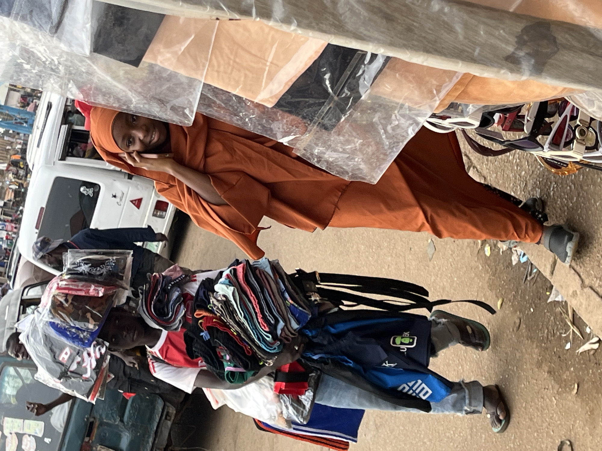 In the city of Bukuru, Nigeria, a young person, likely a boy, walks carrying a large stack of folded clothes balanced on their head, all wrapped in plastic bags. They wear a red and white t-shirt and blue jeans, with dark sandals on their feet. In their arms, they hold another bundle of folded clothes. A blue backpack is also slung over their shoulder. To their right, a young person, likely a girl, wearing an orange headscarf and orange garment, is speaking on a mobile phone. They are standing near a display of sunglasses, which are covered in plastic sheeting. The environment appears to be a street or market, with vehicles and other people visible in the background. The ground is paved with rough material, and there is debris scattered around. The scene captures a moment of everyday life in a busy urban setting, with the young people engaged in commerce or daily activities.