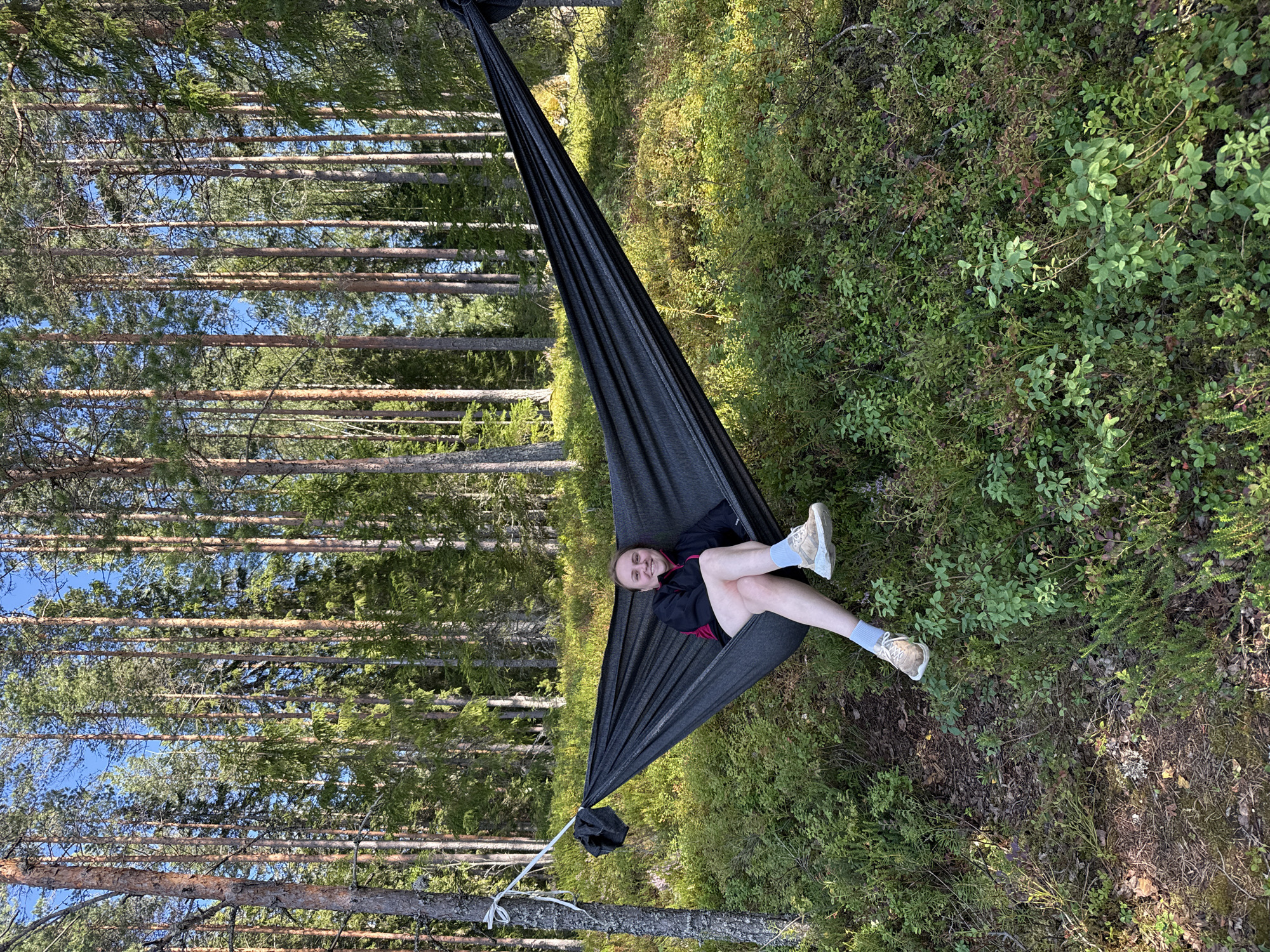 A woman is relaxing in a black hammock strung between two trees in a dense forest. She is smiling and looking at the camera. The hammock is angled downwards from left to right, with the woman positioned in the upper left quadrant of the hammock. She is wearing a black jacket with a pink accent, a short grey skirt, light grey socks, and light brown sneakers. The forest floor is covered in green undergrowth, including bushes and moss. The background consists of tall, slender pine trees with a clear blue sky visible above. The scene suggests a peaceful moment in nature, likely during daylight hours due to the bright lighting and shadows. The location appears to be a natural wooded area, consistent with the forests surrounding Tampere, Finland, possibly within a park or recreational area.