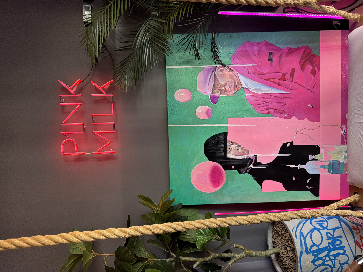 This media file captures a vibrant and modern interior scene, likely within a cafe or shop, located in Khimki, Russia. The composition prominently features a bright red neon sign on a dark grey wall, vertically spelling out "PINK MILK."
Adjacent to the neon sign, a large, colorful painting is displayed. The painting showcases two stylized figures against a background split into vibrant teal green and bright pink sections. The upper figure wears a pink cap, a white undershirt, and a fuchsia jacket, with several pink bubbles floating nearby. The lower figure has a dark, bobbed haircut and a black top, accompanied by a large pink orb and several bottles with liquids. Both painted figures have pale complexions and neutral expressions, depicted in a contemporary art style with abstract elements.
Above the painting, a horizontal purple LED strip light adds to the modern ambiance. The foreground is partially occupied by a thick, braided beige rope, which runs horizontally, suggesting a decorative barrier. Lush green foliage from potted plants is visible both above the rope and near the top of the image, including a palm-like plant and another with broad leaves. One of the planters is white and features blue graffiti-style text and designs. The overall aesthetic is eye-catching and eclectic, combining pop art elements with lush greenery and striking lighting.