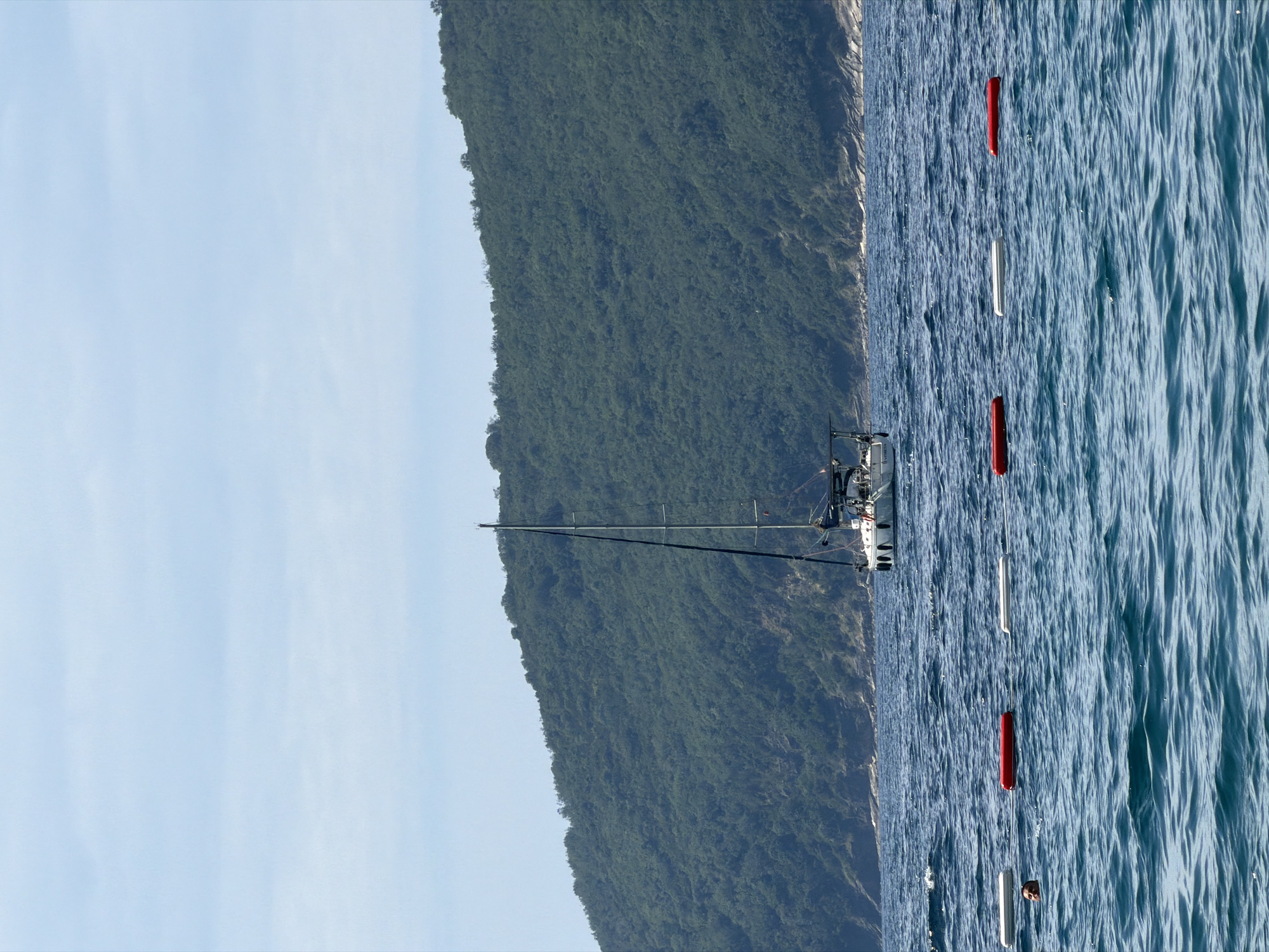 A sailboat is in the sea near Bečići, Montenegro. It is a clear day. A mountain covered with dense green vegetation is visible in the background. There are red and white buoys floating in the water. The sky is blue.