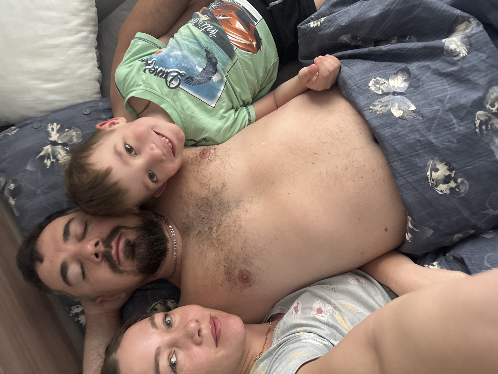 The image shows a family of three lying in bed. A man with a beard and closed eyes lies on his back. A young boy in a green shirt rests his head on the man's chest and smiles at the camera. A woman in a light-colored patterned shirt is positioned below the man, looking at the camera. They are under a blue and gray butterfly-patterned duvet on a bed with blue sheets. A white pillow is visible behind the boy. The image appears to be taken from above, looking down on the family. The location is presumed to be Chaikovsky, Russia.