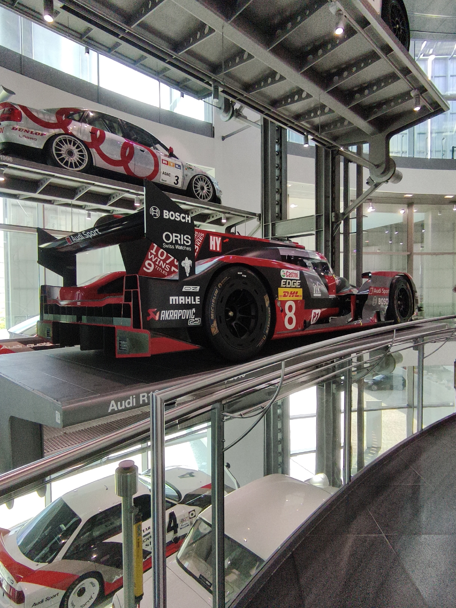 In Ingolstadt, Germany, at the Audi Forum museum, a red and black Audi R18 TDI race car is displayed on a platform. The car features sponsor logos such as Bosch, Oris, and Michelin. Above and to the left of the race car, another Audi race car, white with red and black accents, is suspended. This car displays the Audi Sport logo and the number 3. The museum has a modern, industrial aesthetic with exposed metal structures and large windows.