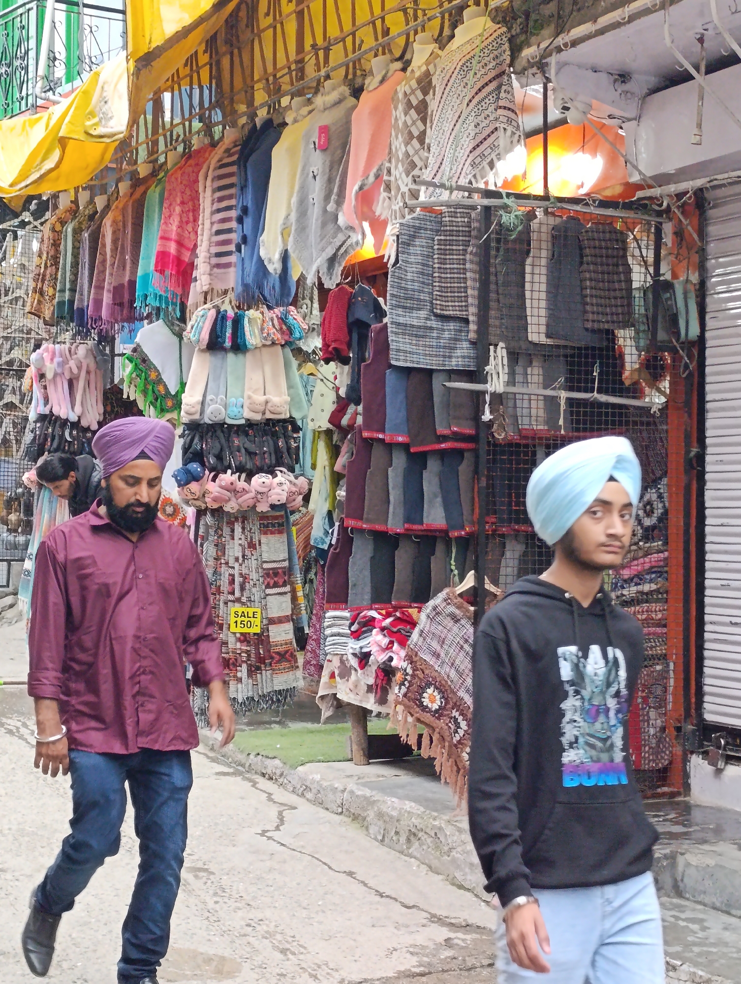 The image depicts a street scene in Vashisht, India, likely a market area given the abundance of goods displayed. In the foreground, two individuals wearing turbans are walking. The person on the left, an adult male with a dark beard, is wearing a maroon button-down shirt and dark blue jeans, accessorized with a silver bracelet. The person on the right, a younger male with a lighter complexion, is wearing a light blue turban and a black hooded sweatshirt with a graphic print. Behind them, a variety of items are displayed for sale, including colorful scarves, sweaters, and what appear to be blankets or shawls, hanging from racks and draped on display. The stalls are partially covered by yellow awnings. The ground is a paved walkway. The overall impression is of a bustling marketplace.