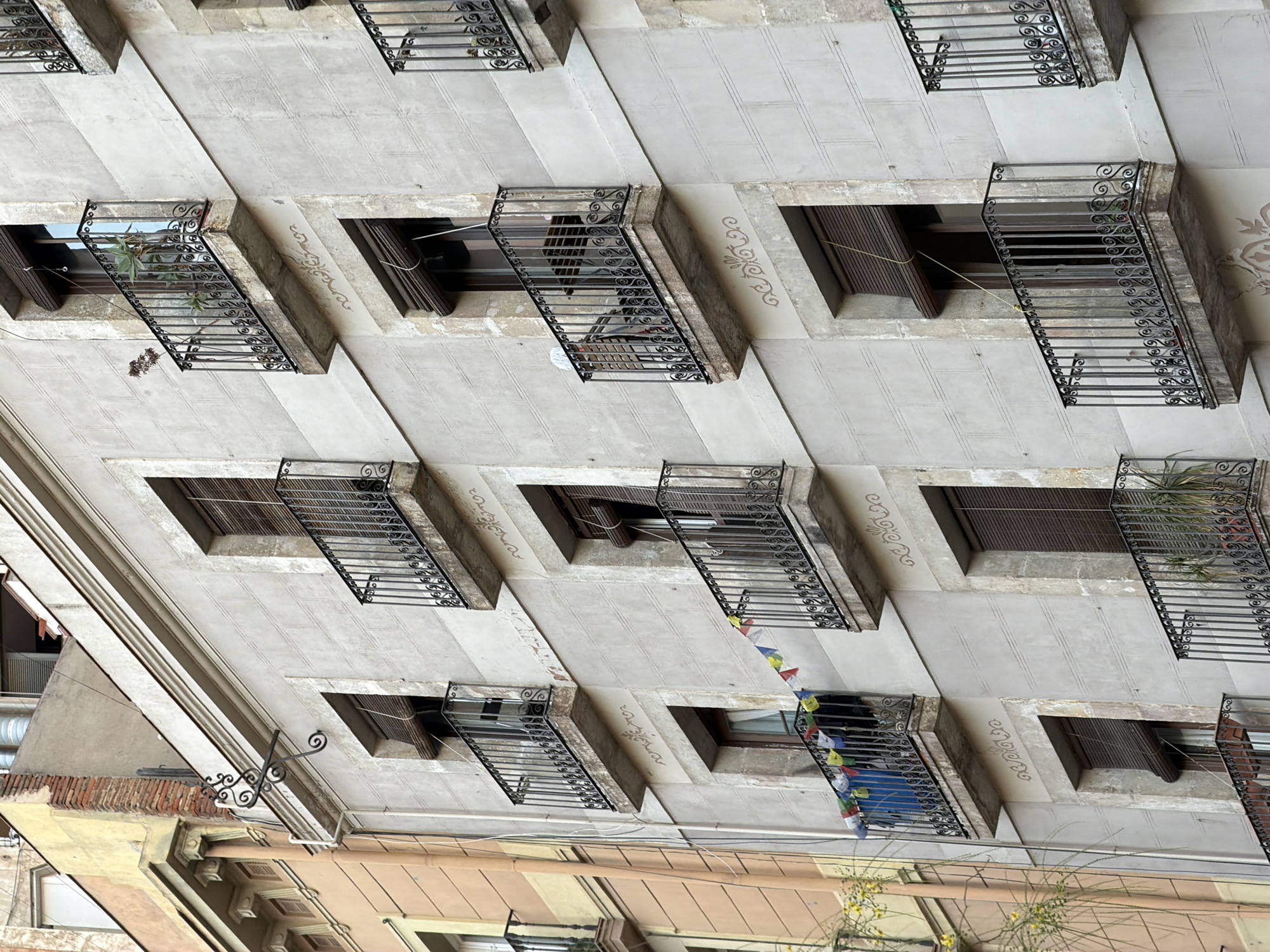This image shows a detail of the facade of a building in Barcelona, Spain. The building has a pale gray stone facade with several balconies featuring wrought-iron railings. The balconies are adorned with plants and some have decorative trim below them. There are also windows with closed blinds. To the left of the building, a portion of an adjacent building with an orange facade is visible. The image is taken from a low angle, looking up at the building.