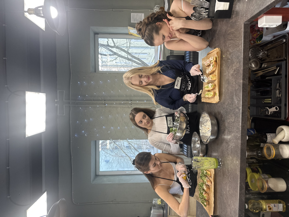 Four adult females are engaged in food preparation at the Mira Kottler Culinary Studio, located at Preobrazhenskaya Square, 7A, Moscow, Russia. The scene is set in a kitchen with a dark grey countertop, light grey walls, and two large rectangular windows in the background revealing an urban exterior with bare trees. Overhead, industrial lighting fixtures and decorative string lights are visible.
From left to right:
1. A female with dark hair, wearing a white spaghetti-strap top, a black apron, and white gloves, is squeezing a lemon over a wooden board containing multiple bruschetta-style appetizers topped with green herbs. A glass of green liquid is positioned nearby.
2. A female with brown hair, dressed in a light-colored long-sleeved shirt, a black apron labeled "Лиза," and white gloves, holds a metal bowl containing green leafy ingredients and observes the first female.
3. A blonde female, wearing a dark blue pinstriped collared shirt, a black apron labeled "Настя" and "МИРА КОТТЛЕР," uses a spoon to apply a white creamy topping to appetizers arranged on a second wooden board. She wears white gloves.
4. A female with dark brown hair in a high ponytail, wearing a white spaghetti-strap top and a black apron, smiles while observing the prepared food, resting her chin on her left hand.
Additional items on the countertop include empty metal bowls. The lower section of the image reveals an open shelf or drawer system containing various cooking oils, sugar or salt containers, a mortar and pestle, and an organized utensil tray. The activity appears to be a collaborative cooking session.