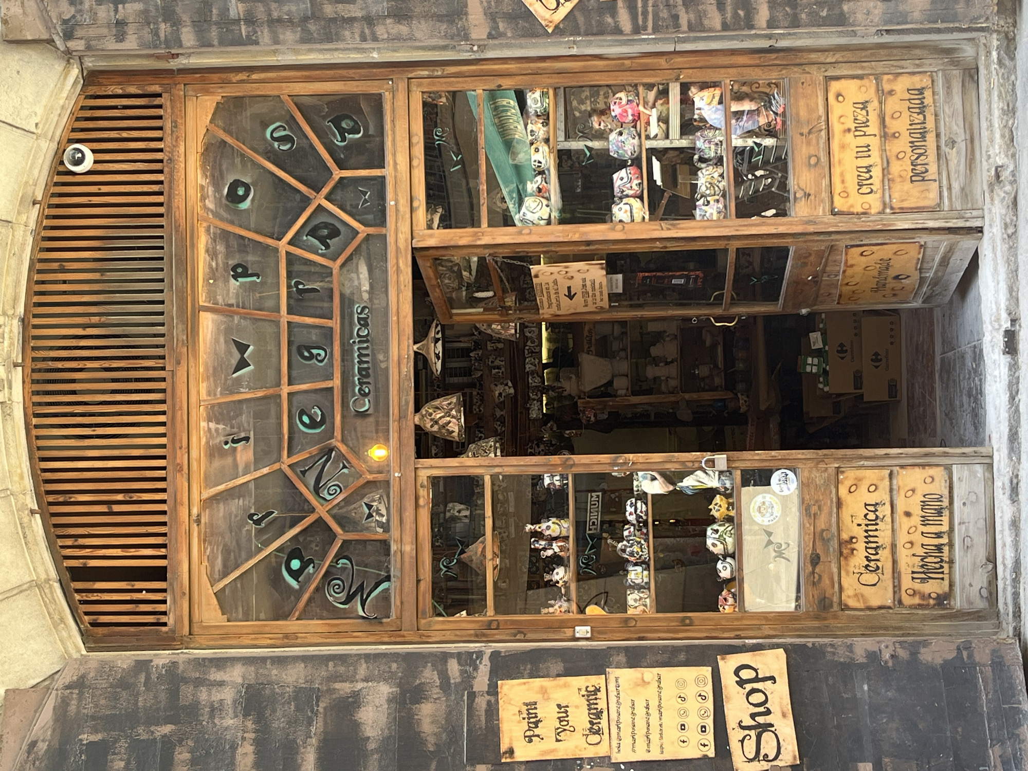 This image captures the rustic storefront of a ceramics shop named "MARIPOSAS CERAMICAS" (Butterflies Ceramics) in Barcelona, Spain. The shop features a large wooden door and glass windows, divided by a distinctive, multi-faceted wooden frame. Inside, various colorful and intricately designed ceramic items, some resembling decorative skulls or pots, are displayed on shelves.
Several hand-painted wooden signs are visible on and around the storefront, mostly in Spanish. On the right side of the door, signs read "crea tu pieza" (create your piece) and "personalizada" (personalized), suggesting customization options. Below these, "Ceramica" and "Hecha a mano" (Handmade) further emphasize the nature of the products. Other signs include "Pedidos" (Orders) and "Handmade". On the left, an English sign invites customers to "Paint Your Ceramic," alongside a "Shop" sign and social media handles like "@mariposamegrabar." A small, white security camera is mounted on the wooden slatted archway above the left side of the door. The overall impression is that of a charming, artisanal small business. The daylight indicates it's daytime, and reflections on the glass suggest the store is open for business.
