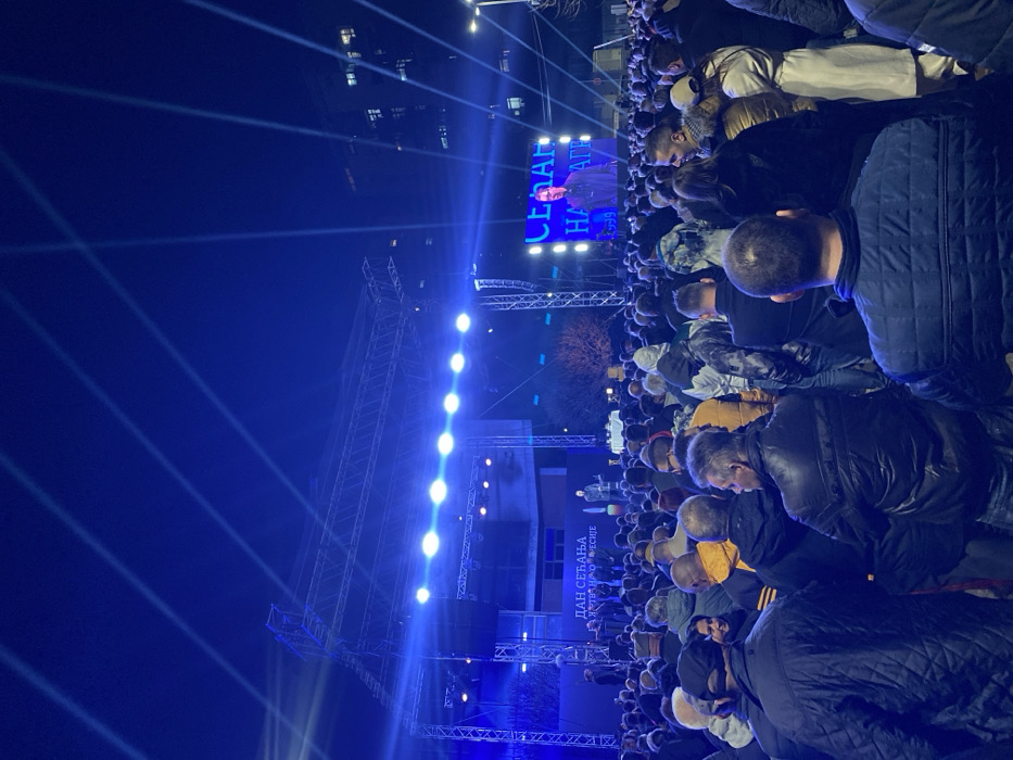 The image depicts a nighttime outdoor public gathering in a city square in Vranje, Serbia. A large crowd, estimated in the hundreds, primarily adults, occupies the foreground and midground, facing a brightly lit stage and an adjacent large LED screen.
The central feature is an elevated stage constructed from a metal truss system, illuminated intensely by numerous blue floodlights and spotlights. These lights cast prominent blue beams upwards into the dark, clear night sky. On the stage backdrop, text in Cyrillic reads "ДАН СЕЋАЊА ЖРТВАМА НАТО АГРЕСИЈЕ" (DAY OF REMEMBRANCE FOR VICTIMS OF NATO AGGRESSION). A speaker is visible on the stage, addressing the assembly.
To the right of the stage, a large rectangular LED screen displays a close-up image of a man speaking, with Cyrillic text visible, including "СЕЋАЊЕ НА ДРАГЕ..." (REMEMBERING DEAR...). The crowd is densely packed, with individuals generally standing and directing their attention towards the stage and screen. People are dressed in various casual to semi-casual outerwear, consistent with an evening outdoor event.
In the background, behind the stage and to the sides, multiple-story urban buildings with illuminated windows are visible. Bare trees or shrubs are discernible behind the stage structure. The overall lighting scheme is dominated by the blue artificial light from the stage.