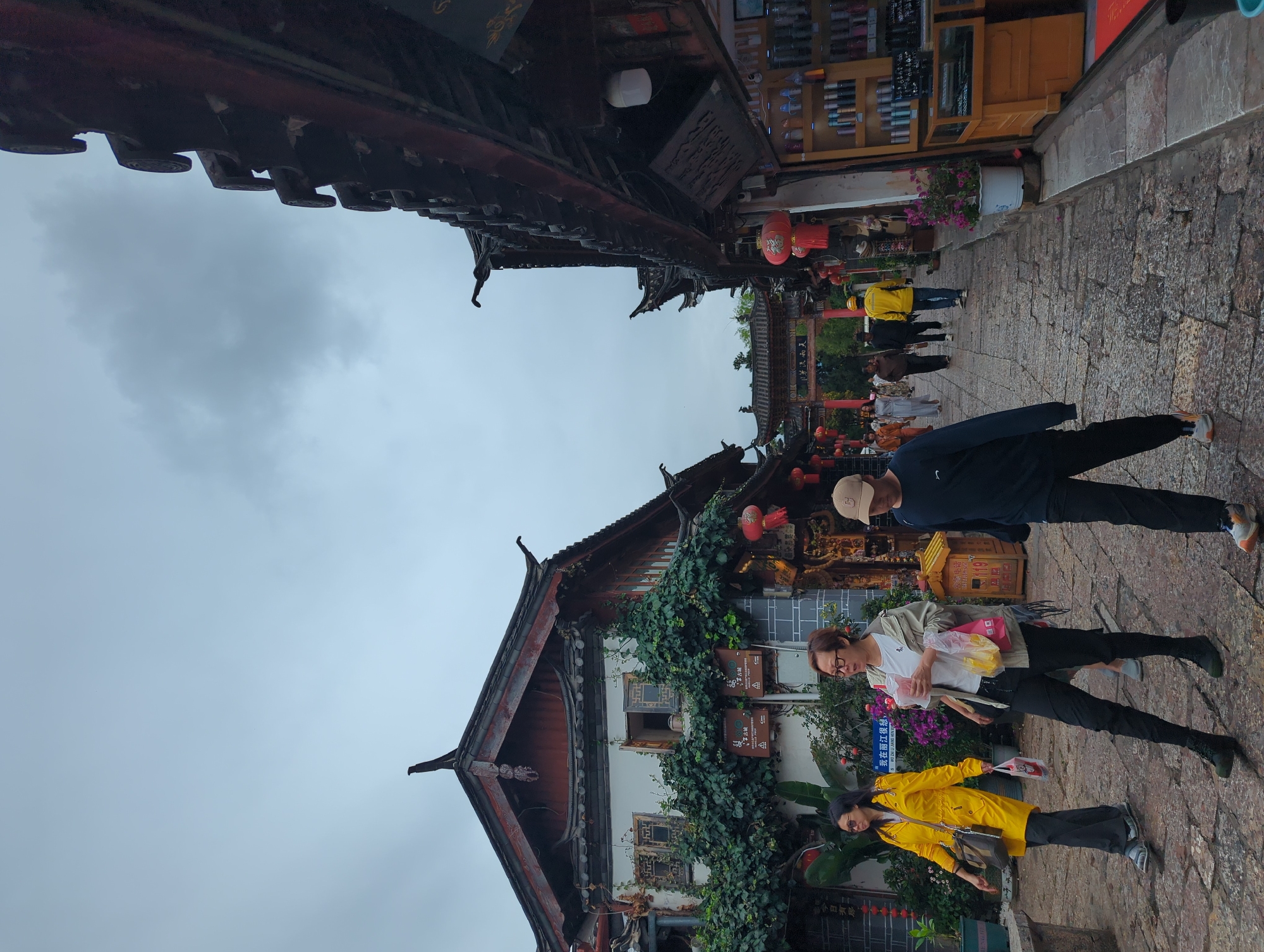 The image shows a street scene in the Gucheng District of China. People are walking on a stone-paved street lined with traditional Chinese architecture. The buildings have dark tiled roofs and wooden facades, with some adorned with greenery. Red lanterns hang from some of the buildings, suggesting a festive atmosphere or the presence of shops.
Several individuals are visible in the foreground and background. One woman in a yellow jacket is walking and holding a newspaper. Another woman in a lighter jacket is carrying a bag and appears to be looking at something. A man in a dark long-sleeved shirt is walking behind them. Further down the street, other people are seen walking, including one person in a yellow shirt and a person in a blue shirt.
The sky is overcast, indicating a cloudy day. The overall setting appears to be a pedestrian area within a historic town or district. Some signage with Chinese characters is visible on the buildings, though the text is not clearly legible in this image. The scene suggests a leisurely stroll through a culturally rich environment.