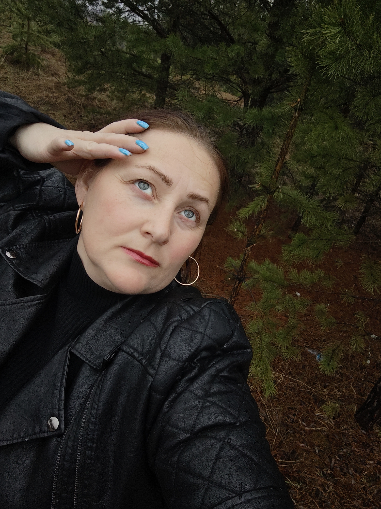 A medium-close-up, eye-level selfie shows a woman outdoors in what appears to be a natural, wooded setting. The woman, with fair skin and light reddish-brown hair, is looking upwards and slightly to her right, with her left hand raised to her forehead, fingers spread, as if shielding her eyes or shading herself. Her eyes are blue, and she wears red lipstick.
She is dressed in a black quilted leather jacket over a black turtleneck. Gold hoop earrings are visible on both ears. Her fingernails are painted a vibrant sky blue. The jacket appears to have some moisture on it, suggesting damp weather or recent rain.
The background consists of dense coniferous trees, primarily pine trees with dark green needles. The ground cover is a mix of reddish-brown pine needles and dry foliage, with some sparse green undergrowth. The lighting suggests an overcast or muted daytime setting. No text is visible in the image. The overall impression is one of a contemplative moment in a damp, forested environment in Chaikovsky, Russia.
