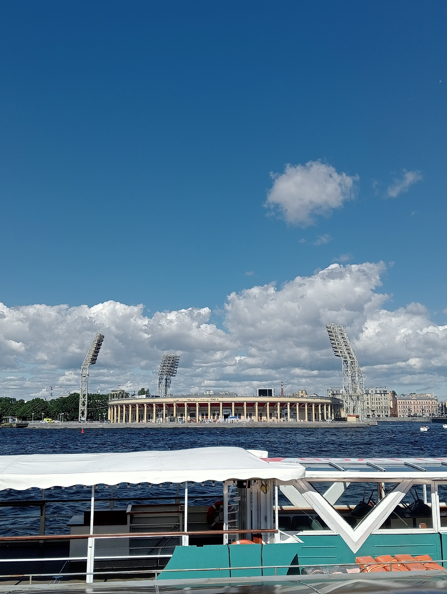 This is an outdoor, eye-level shot of a body of water with a stadium in the background and a boat in the foreground. The stadium is a large, circular structure with columns and multiple tiers. There are three large floodlight towers positioned behind the stadium. The sky is blue with scattered white clouds. The water is choppy, indicating a moderate breeze. The boat in the foreground is white with a teal hull and has a white canopy. Life vests are visible on the side of the boat. The stadium is likely Petrovsky Stadium, located on Petrovsky Island in Saint Petersburg, Russia.