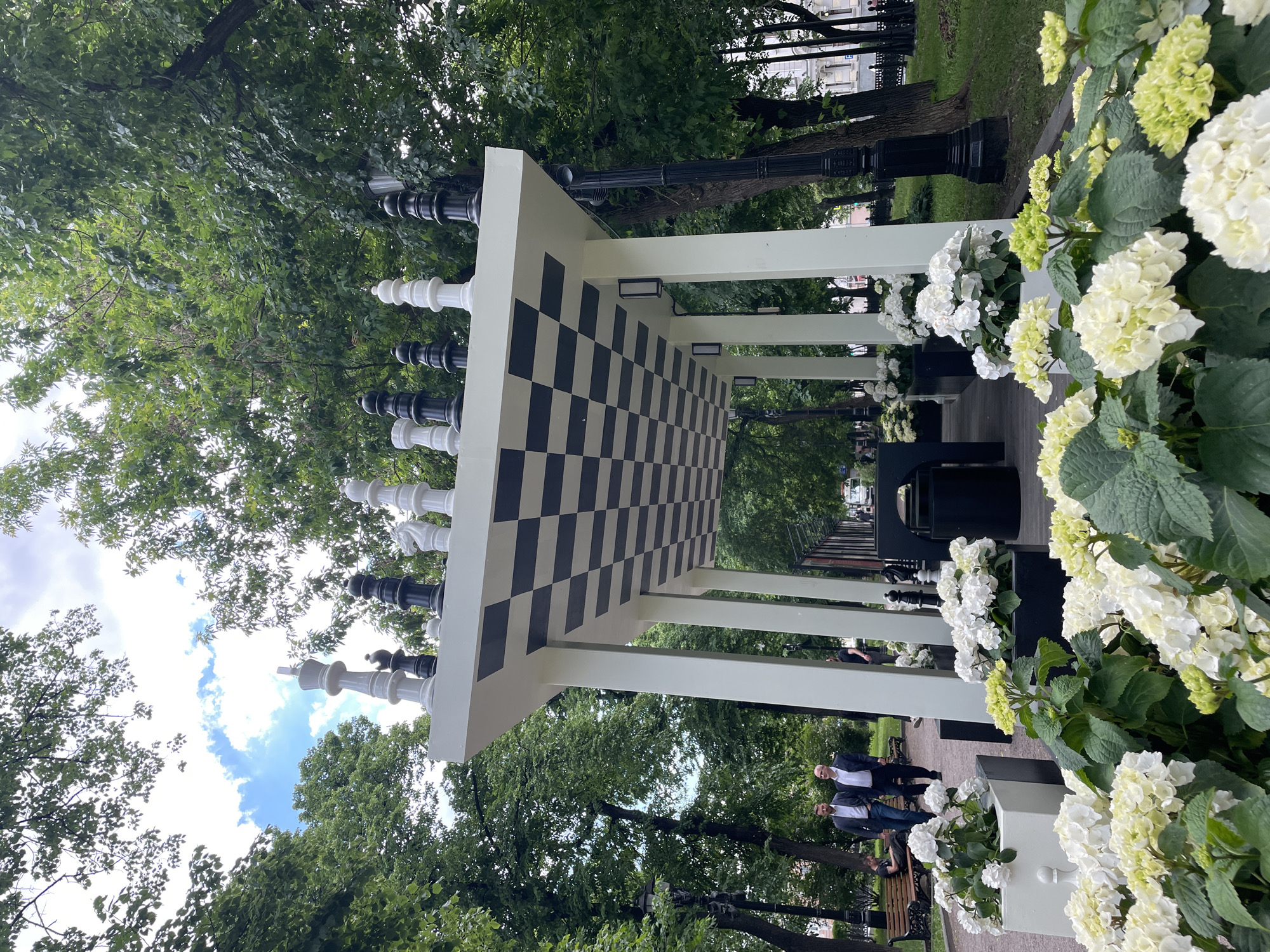 This media file depicts a vibrant public park or garden in Moscow, Russia, centered around a striking architectural feature: a long, white pergola-like structure with a giant chessboard pattern on its underside. Large, decorative chess pieces in black and white (kings, queens, knights, and pawns) are arranged along the top edges of this canopy.
The setting is lush and green, with numerous mature trees providing ample foliage and shade. Beneath the structure and along its sides, planters are filled with abundant white and light green hydrangeas, adding bursts of color and a cultivated garden feel. Traditional black streetlights and park benches are visible further down the pathway, nestled among the trees.
Several people are present, indicating a casual, leisurely atmosphere. Two men in dark jackets and light shirts are standing and conversing near the middle right of the image. Other individuals can be seen walking or seated on benches in the background, enjoying the outdoor environment.
The time of day appears to be daytime, with natural light suggesting either late morning or afternoon. The sky is partly cloudy, with visible patches of blue, indicating pleasant weather suitable for outdoor activities. No discernible text is visible within the frame. The overall scene conveys a sense of tranquility and public enjoyment in a well-maintained urban green space.