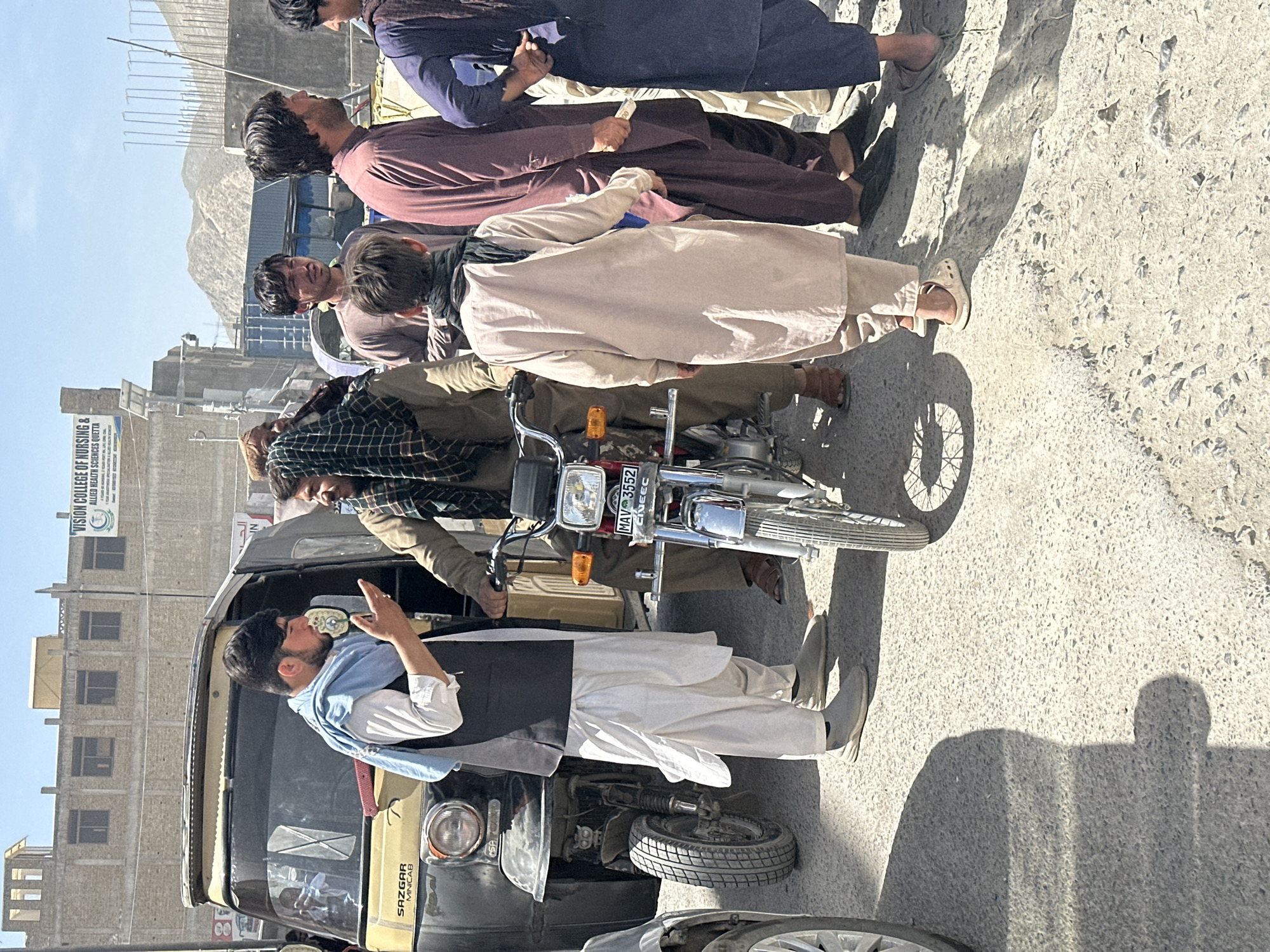 The image depicts a vibrant street scene under bright daylight, featuring several men and vehicles. In the immediate foreground, a three-wheeled vehicle, similar to an auto-rickshaw or minivan, is visible. A man, dressed in a white kurta, black vest, and a light blue shawl, leans out of its passenger side, looking forward, with his right hand raised near his face, possibly engaged in a conversation or holding an object. The vehicle has "SAZGAR MINICAB" printed on its side.
Adjacent to this vehicle, a red and chrome motorcycle is present, displaying the license plate "MAV 5552". A man in a plaid shirt and brown trousers stands next to the motorcycle, holding its handlebars. Behind him, several men are gathered, standing in a line. They wear traditional attire including kurtas in beige, maroon, and dark blue. Their distinct, long shadows cast on the asphalt road indicate strong overhead sunlight, suggesting it is likely midday or early afternoon.
In the background, buildings line the street. A prominent sign on one building is clearly visible, reading "COLLEGE OF NURSING & ALLIED HEALTH SCIENCES QUETTA" and partially "VISION COLLEGE OF...". Beyond the urban structures, rugged mountains are visible under a clear, blue sky, placing the scene in a location like Quetta, Pakistan. The overall atmosphere suggests a typical day with people going about their routines in a sunny, bustling environment.