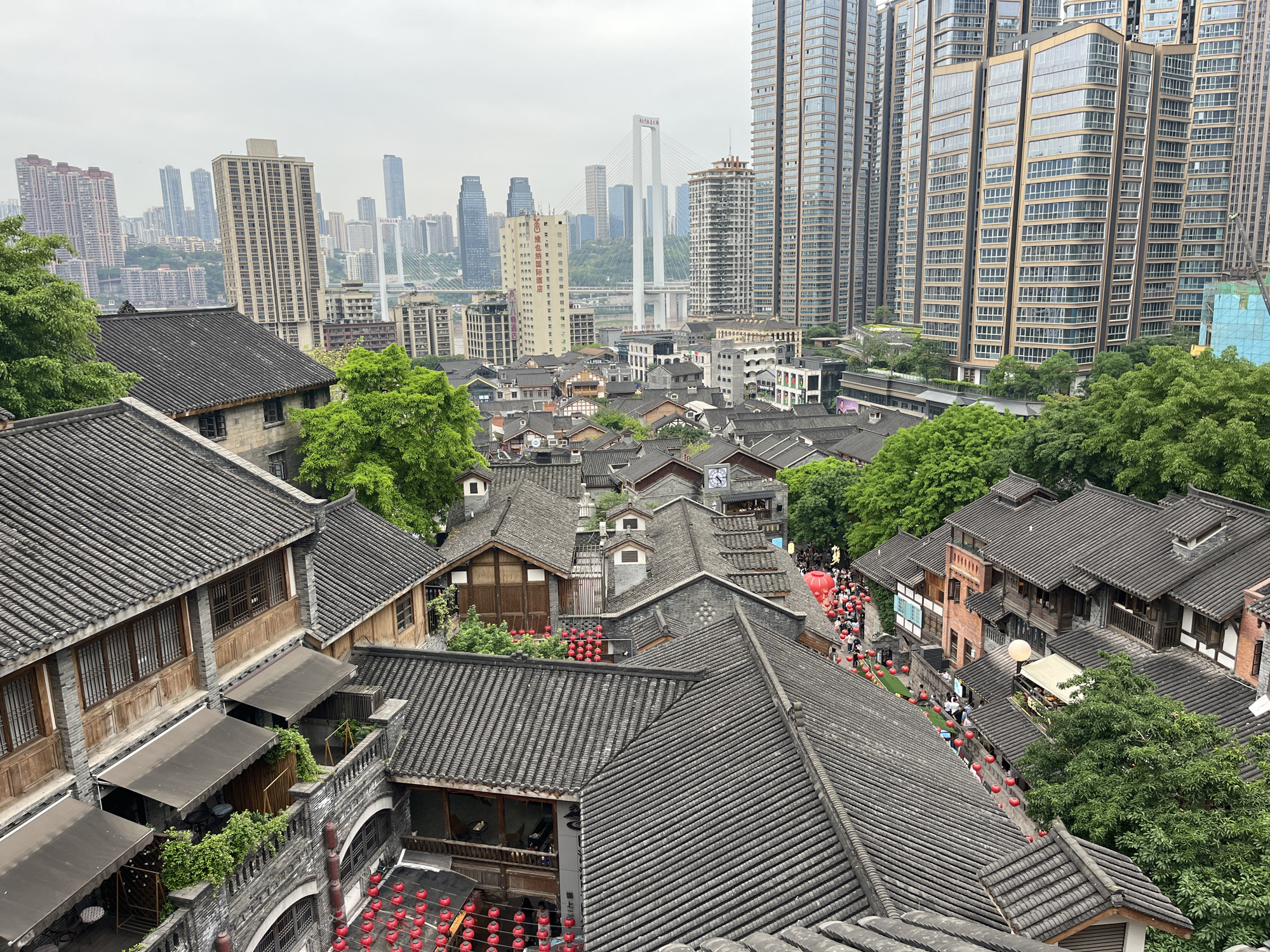 The image captures a vibrant panoramic view showcasing a striking contrast between traditional Chinese architecture and a modern urban skyline in Yuzhong District, China. The scene is set under an overcast sky during daytime, providing diffused light.
In the foreground and midground, a dense cluster of multi-story buildings with distinctive gray tiled roofs, intricate wooden facades, and balconies dominates the lower half of the frame. Lush green trees and foliage are interspersed among these structures. Numerous red lanterns are prominently displayed, strung across pathways, hanging from eaves, and adorning outdoor seating areas, creating a festive and inviting atmosphere. Many people are seen strolling along the winding pedestrian paths and streets within this traditional district, suggesting a lively market or tourist area. A large red balloon or float is also visible among the crowds, and a clock face can be seen on one of the buildings.
Beyond this historical enclave, the background is defined by a sprawling, dense modern cityscape. Numerous towering skyscrapers, primarily residential and commercial buildings with glass and concrete facades, rise dramatically. A prominent white cable-stayed bridge with two tall pylons spans a distant waterway, a distinctive landmark connecting parts of the city. Other high-rise buildings and urban infrastructure stretch across the horizon.
Visible text includes Chinese characters, notably "维也纳国际酒店" (Vienna International Hotel) on a tall building in the mid-distance. The overall image captures the dynamic coexistence of historical heritage and contemporary development characteristic of a major Chinese city.