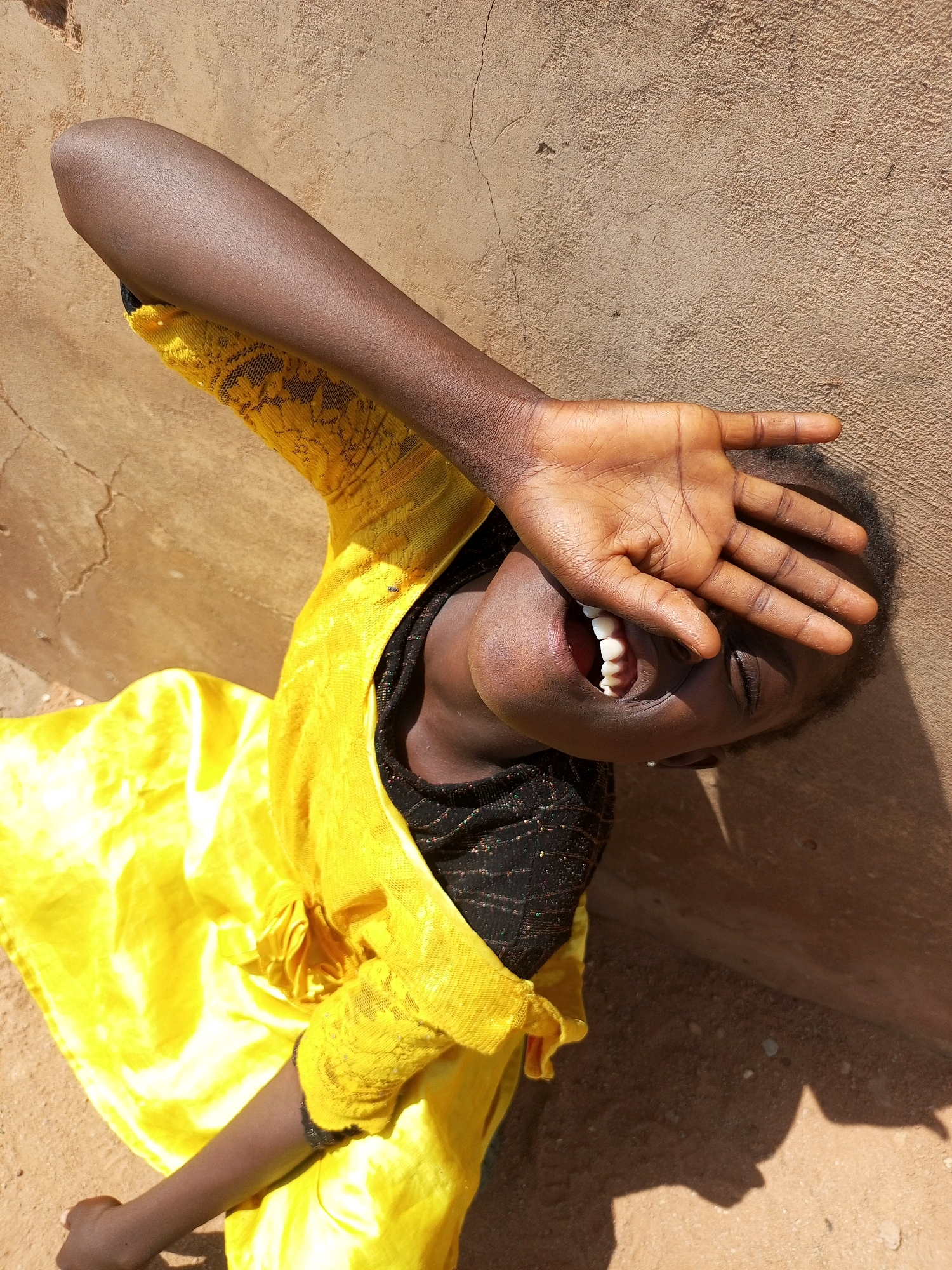 A young person, possibly a child, is lying down and laughing outdoors against a textured, tan-colored wall. They are wearing a bright yellow garment over a dark, possibly black, inner garment. The person's right arm is raised, covering their face from the eyes down, with their hand spread open. Their mouth is wide open, revealing teeth, and their eyes are closed. The ground beneath them appears to be dirt or sand. The lighting suggests it is a sunny day, casting a shadow of the person on the ground. The specific location in Tempure, Nigeria, cannot be determined from the visual information provided.