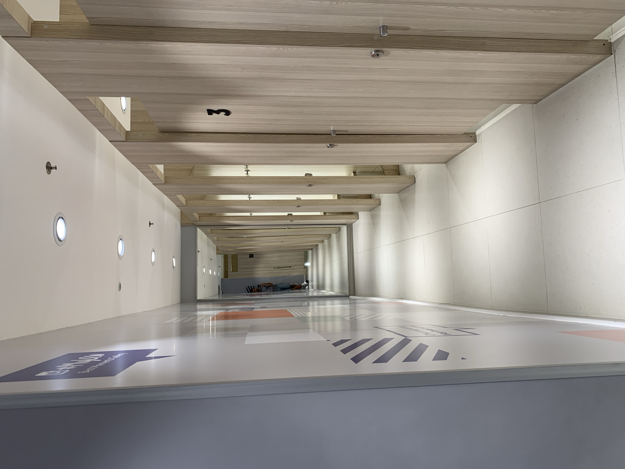 The image shows a long hallway with light-colored wooden slats on the ceiling. The walls are white, and there are round lights embedded in the left wall. The floor is light gray with arrow markings and text. In the distance, at the end of the hallway, there is a mirror reflecting the scene, including people lying on the floor. The location is likely an interior space, potentially a gallery or museum, in an unknown city, as information about Porlamar, Venezuela is not evident.