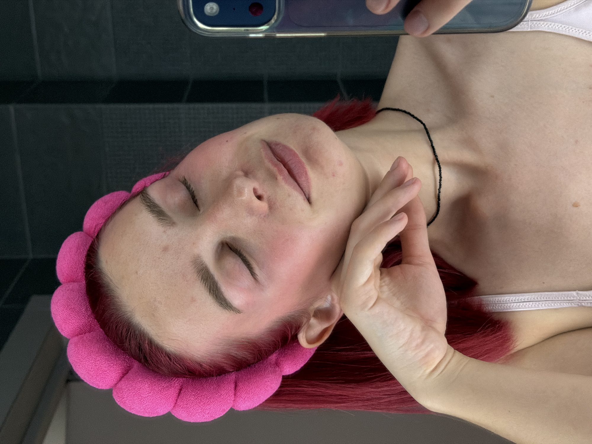 A young woman with bright pink hair and closed eyes is taking a selfie in a dimly lit bathroom. She is wearing a fluffy pink headband and a white tank top. Her left hand is resting on her cheek, and she is looking at her phone, which is held up to take the picture. The background is a dark tiled wall, and a mirror is visible to the left of the woman, reflecting her image. The scene appears to be domestic and personal, suggesting a moment of self-care or a casual snapshot. There are no indications of the location being specifically in Chaikovsky, Russia, or any other specific city.