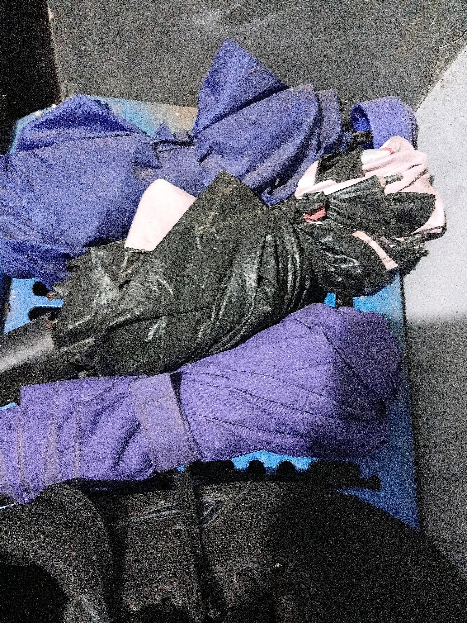 The image shows a close-up, overhead view of a pile of laundry and a shoe, situated on a blue plastic surface, possibly a pallet or crate. The laundry consists of several items, predominantly in shades of purple and black, with some pink fabric visible as well. One prominent item is a rolled-up purple garment, possibly an umbrella or a long piece of fabric, secured with a strap. Other items are loosely piled and appear to be fabric. In the foreground, a black shoe with laces is visible. The background is a dark, textured surface, possibly a wall or floor, with a lighter gray area to the right. There are no people visible in the image. The scene appears to be indoors or in a sheltered outdoor area, given the artificial lighting and the presence of plastic material. Without further context or identifiable landmarks, it is impossible to determine the specific location in Cikal, Indonesia.