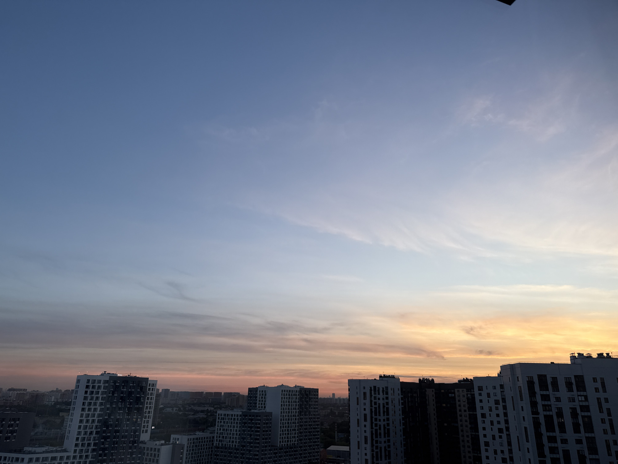 The image depicts a cityscape at sunset, as seen from a high vantage point. The sky transitions from a clear, soft blue at the top to a gradient of pink, orange, and yellow hues near the horizon, indicating the setting sun. Wispy clouds are scattered across the sky, catching the warm light.
Dominating the lower half of the frame is a collection of modern high-rise residential buildings. These buildings are characterized by their dark and white facades, with numerous windows visible. The architectural style appears uniform, suggesting a planned residential development. Some buildings are more sharply defined, while others are partially obscured by distance or shadow.
In the far distance, a denser collection of buildings forms a hazy skyline, characteristic of a large urban area. The overall impression is one of a densely populated residential district within a city. The image is a still, static shot, with no visible human activity or movement. The environment is clearly urban, with the natural phenomenon of sunset providing the sole dynamic element. Based on the architectural style and the context of Lyubertsy, Russia, this scene is likely a view of a newly developed residential area in or around the city.