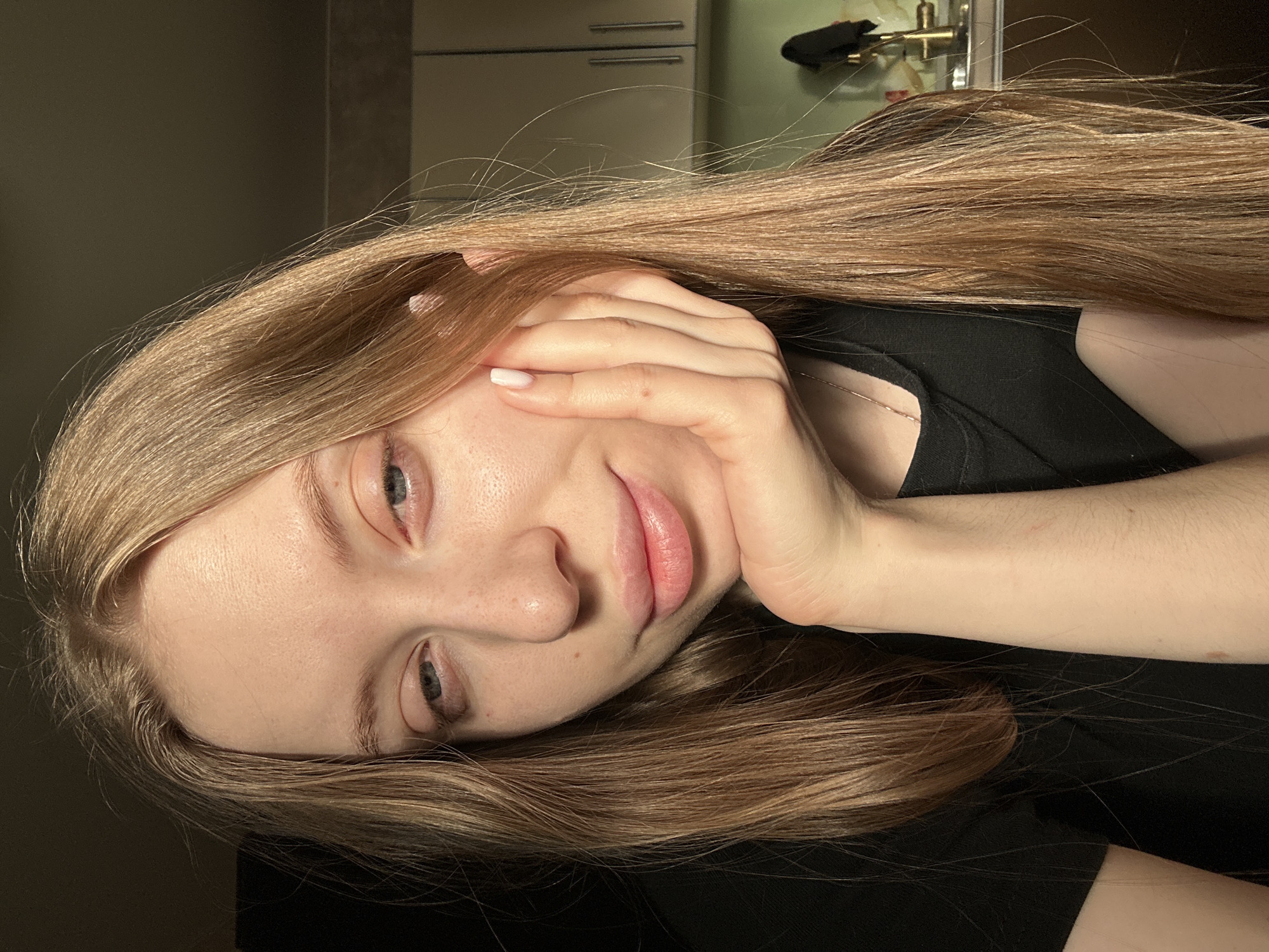 A young woman with long, blonde hair is resting her head on her hand. She is wearing a black top. The background appears to be an interior setting, possibly a kitchen or living area, with cabinets and other household objects visible. There is no information available in the image to verify the location in the city of Chaikovsky, Russia.