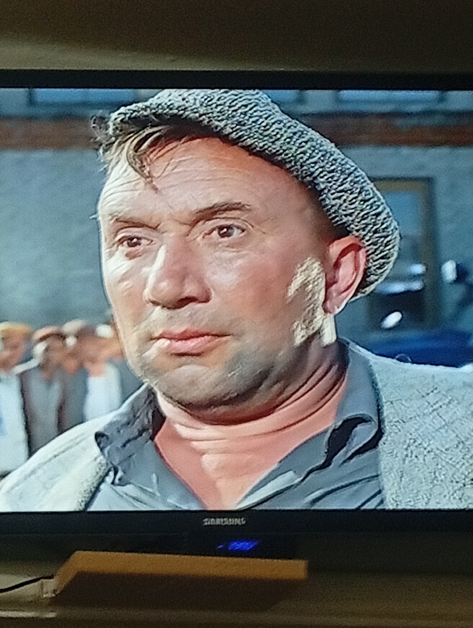 The image shows a close-up, head-and-shoulders shot of a man wearing a patterned flat cap, a dark collared shirt, and a textured gray jacket. He is looking slightly to his left with a neutral expression. The background is blurred, showing what appears to be a building with windows and several out-of-focus figures. The man's skin is weathered, and he has wrinkles around his eyes and forehead. The lighting suggests an indoor or overcast outdoor setting. There is no visible indication of the city of Pushkino, Russia, in this image.