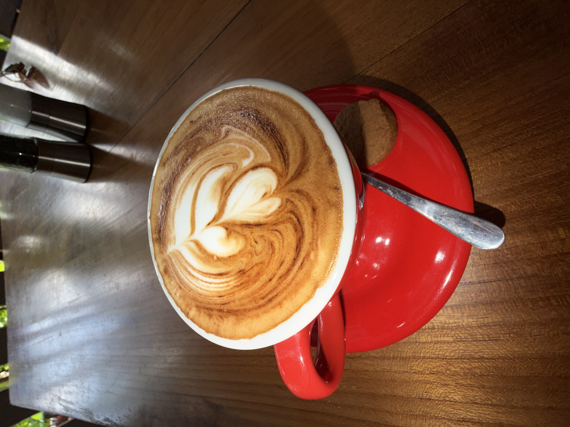 A top-down, medium shot features a cappuccino in a white-rimmed red mug, resting on a red saucer with a spoon and a small cookie. The cappuccino has latte art in the shape of a heart or flower. Two metallic containers are on the table to the upper left. The image is captured on a wooden table with visible wood grain. The presence of bright spots on the table and reflections off the containers suggests it is outdoors or near a bright window, possibly in North Kuta, Indonesia.