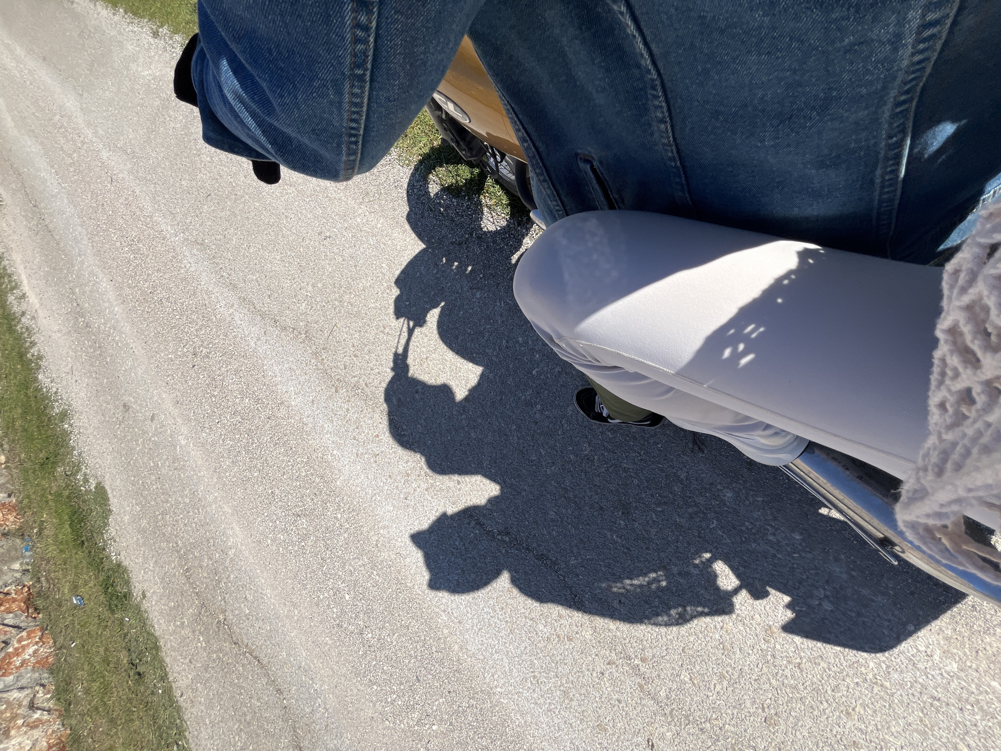 The image presents a first-person perspective from atop a motorcycle, facing the front. The motorcycle is positioned on an asphalt road. A person wearing a blue denim jacket and light-colored pants is seen straddling the motorcycle. The shadow of the motorcycle and rider is cast onto the road. The edge of the road is visible, with grassy vegetation alongside it. The image captures the view while traveling on a road in an outdoor environment in the city of Kovači, Montenegro.