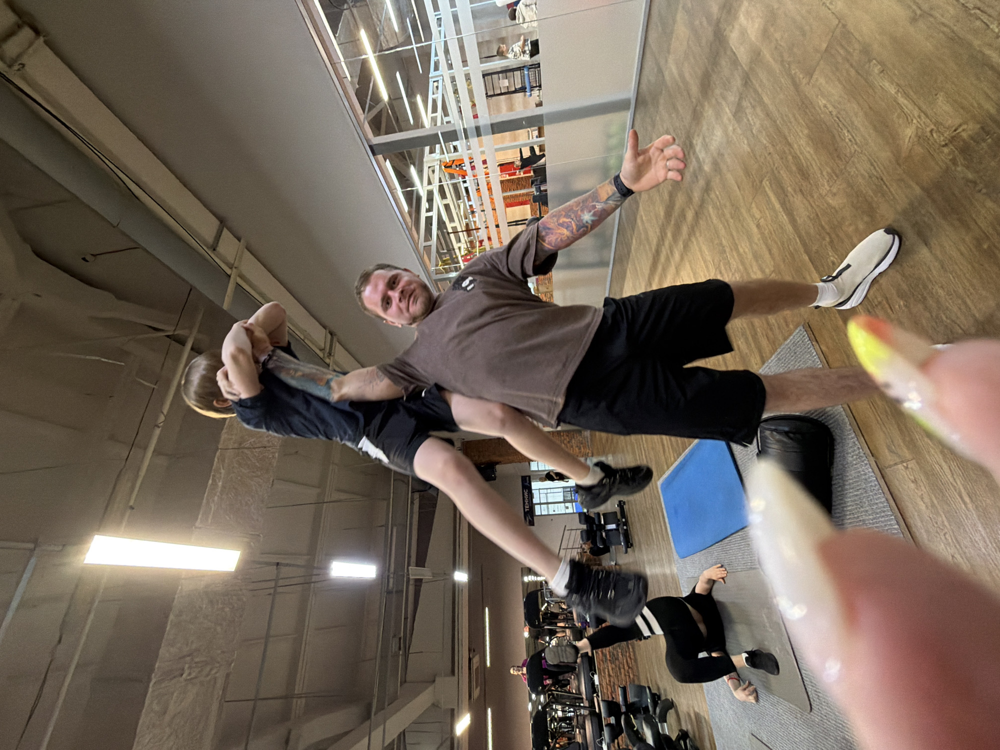 The image appears to be taken in an indoor fitness center. A man is holding another person around their torso, lifting them off the ground. The man is wearing a brown t-shirt and black shorts, with visible tattoos on his arms. The person being held is wearing a dark shirt and shorts.
In the background, other people are using exercise equipment. There are multiple treadmills and exercise bikes visible. A woman wearing black and white striped leggings is sitting on a mat on the floor.
The flooring is wood-like. Exercise mats and a blue square are on the floor. The indoor space appears to be modern with large windows. The location of the city is unknown.