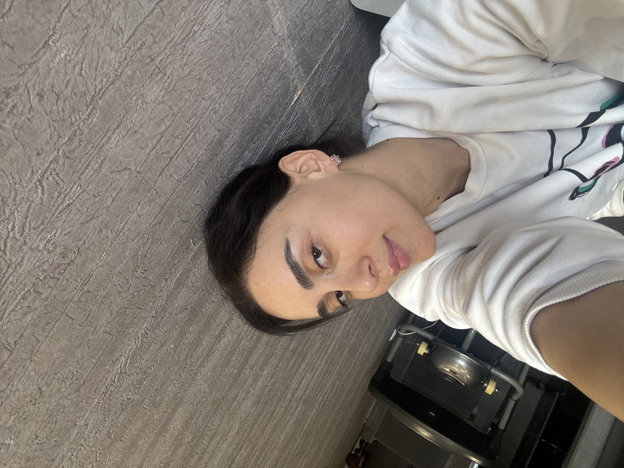 The image features a person, likely a young woman, lying down and looking towards the viewer with a soft, slightly playful smile, her tongue subtly visible between her lips. She has dark hair, neatly styled away from her face, and distinctive, well-defined eyebrows. She is wearing a white crew-neck sweatshirt that has a graphic design on its front, characterized by prominent black lines and hints of teal and pink colors. A small, light pink, possibly flower-shaped, stud earring is visible in her right ear.
The background consists mainly of a textured, grayish-brown surface, resembling a wall or floor, indicating an indoor setting. In the bottom right portion of the image, behind the person's right shoulder, a partial view of a utility unit is visible. This unit includes a metallic bowl or basin, gray piping, and yellow or gold-colored knobs or handles, suggesting it could be part of a sink or a water dispenser. The lighting is even and soft. The overall expression of the person is relaxed and pleasant. No discernible text is visible within the image. The location context for this image is Tbilisi, Georgia.