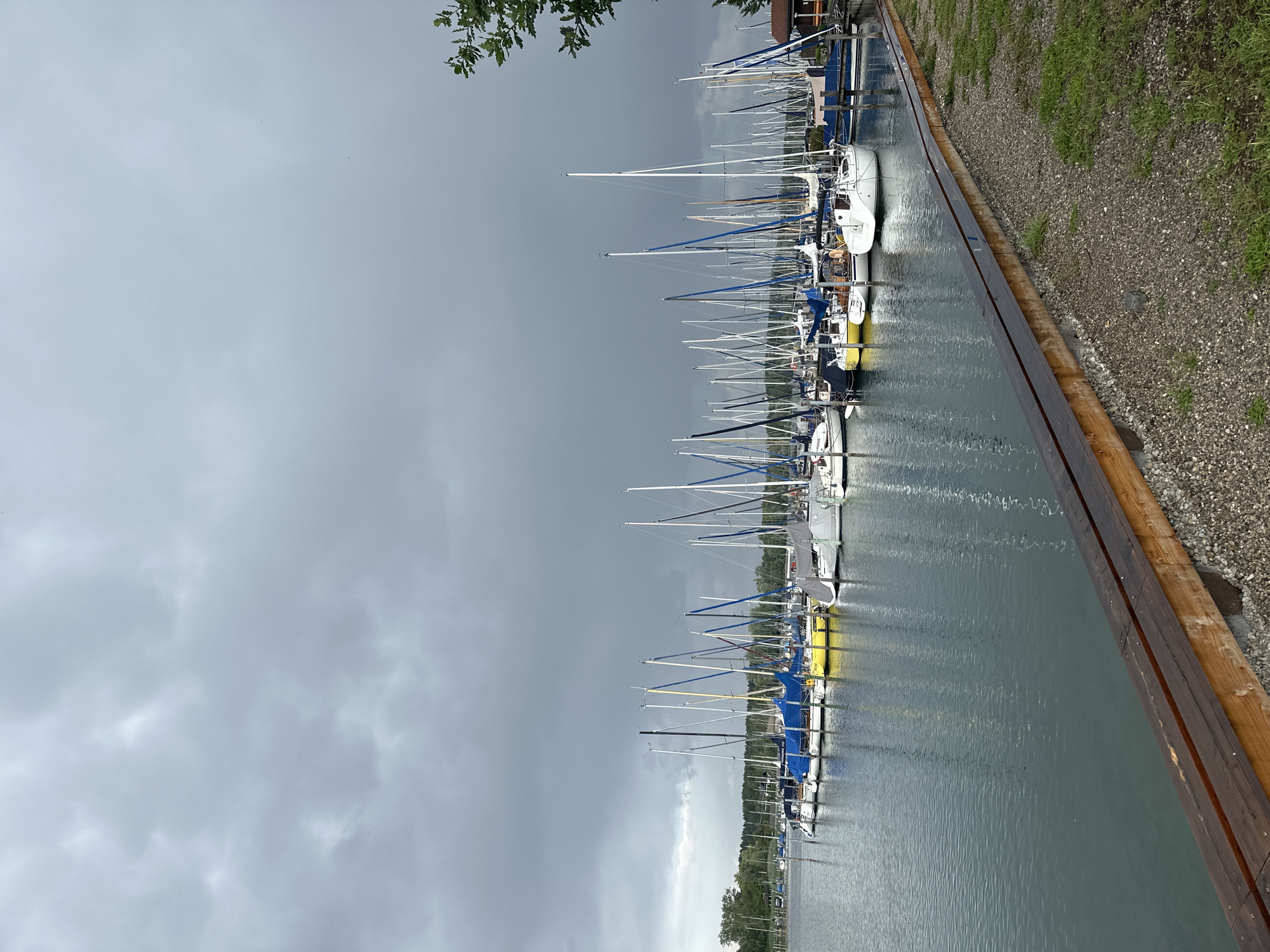 A marina is depicted under a dark, cloudy sky in Seeon-Seebruck, Germany. Numerous sailboats are moored in the water, with their masts extending upwards. Some boats are covered with blue or gray tarps, while others are bare. The water is calm and reflects the sky and the boats. A wooden walkway runs along the edge of the marina, with gravel and grass on the landside. Trees and buildings are visible in the background on the far side of the water. The overall impression is one of a quiet, overcast day at a lakeside harbor.