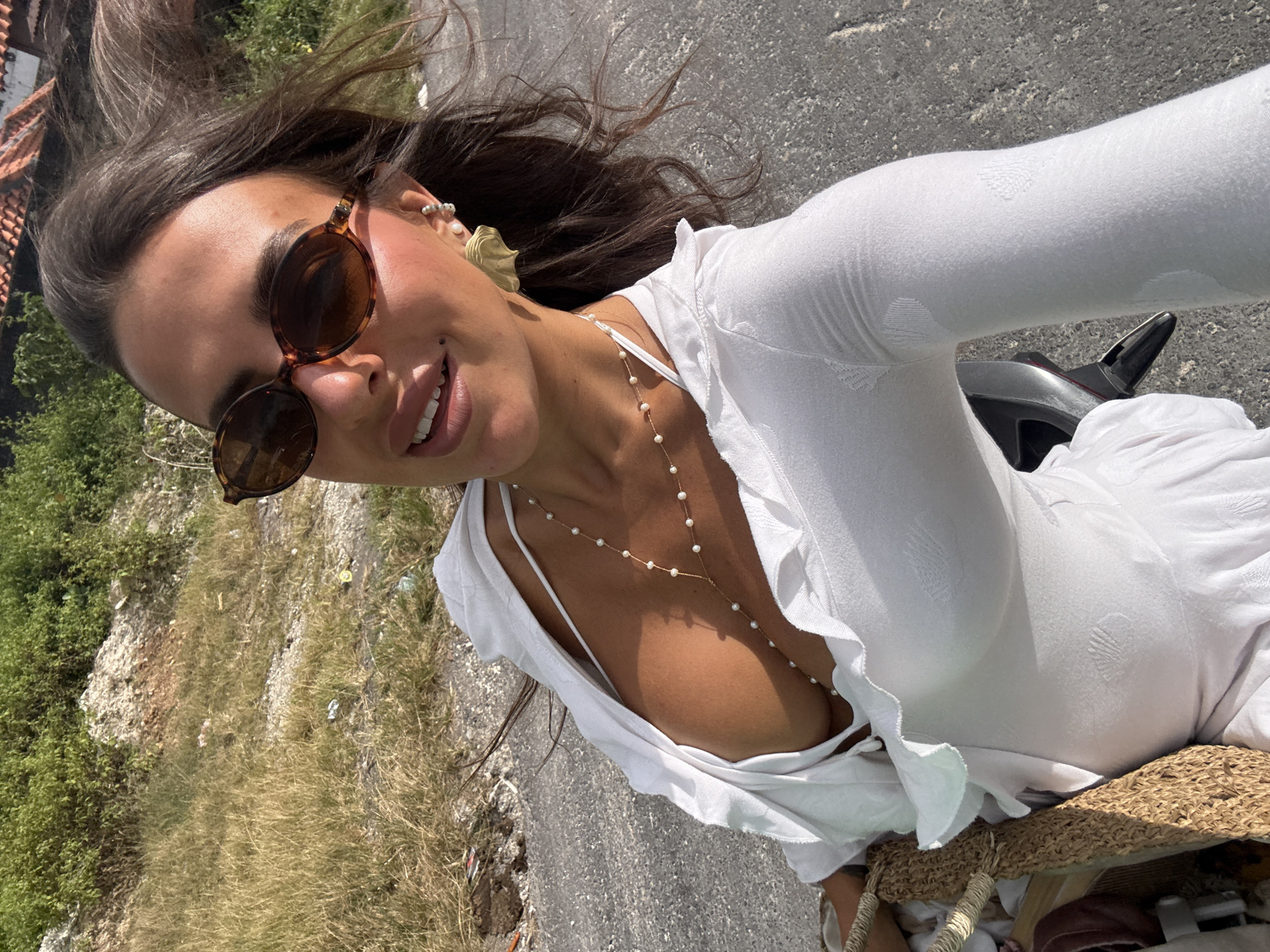 A woman is taking a selfie on a scooter in Kuta Selatan, Indonesia. She is wearing a white dress and sunglasses. The background is a grassy hill with some trees. The road is visible in the foreground.