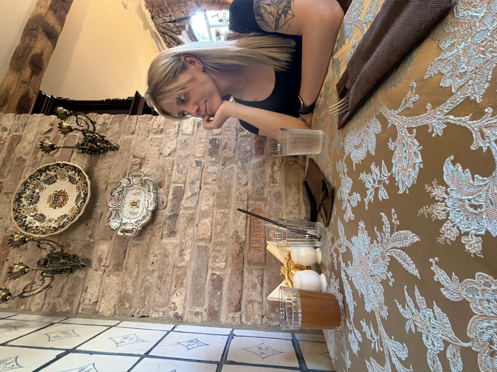 In the city of Suzdal, Russia, a blonde woman with a black shirt and a tattooed arm sits at a table covered with a patterned tablecloth. The table is set with two glasses, one containing a brown beverage, salt and pepper shakers, napkins, and cutlery. The background features a brick wall adorned with decorative plates and a wall sconce. The woman is looking to the side with her hand on her chin, appearing contemplative. The scene suggests a dining experience in a restaurant with a rustic ambiance.