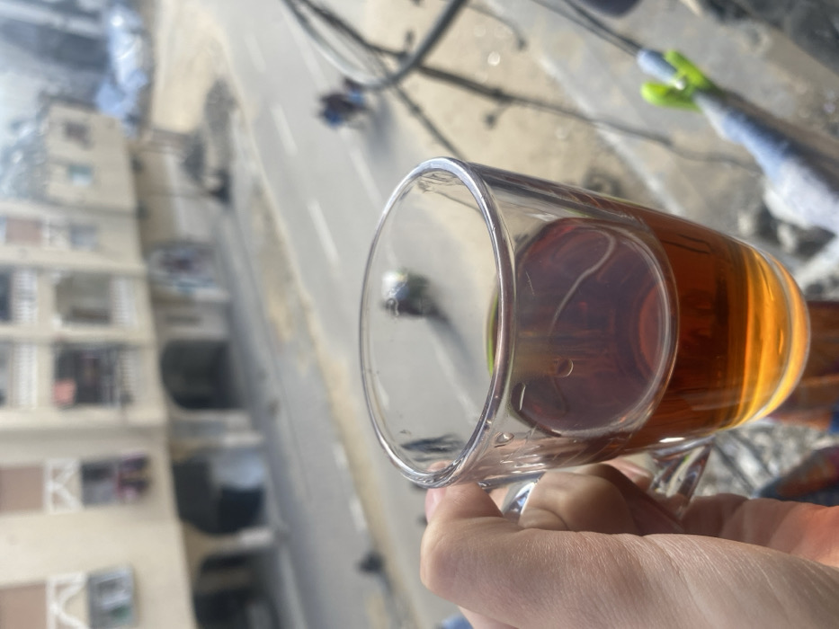 A first-person perspective photograph captures a light-skinned hand holding a clear glass mug containing an amber-colored liquid, consistent with tea, in the foreground. The background, significantly out of focus, depicts an urban environment in Khan Yunis, Palestinian Territory. A two-lane paved road with white dashed center markings is visible in the mid-ground. To the left of the road stands a multi-story building with a light-colored facade, featuring multiple balconies, some displaying hanging laundry, and arched openings on the ground floor. To the right of the road, an area containing piles of debris, some covered by a blue tarp, is present. Several blurred human figures are discernible: two individuals on what appears to be a motorcycle or scooter are on the right side of the road, moving away from the viewer, while other dark, indistinct figures are visible near the debris and further down the road on the left. The scene is illuminated by daylight. Dark, blurred lines, consistent with a railing or overhead wires, are partially visible on the far right edge of the frame.