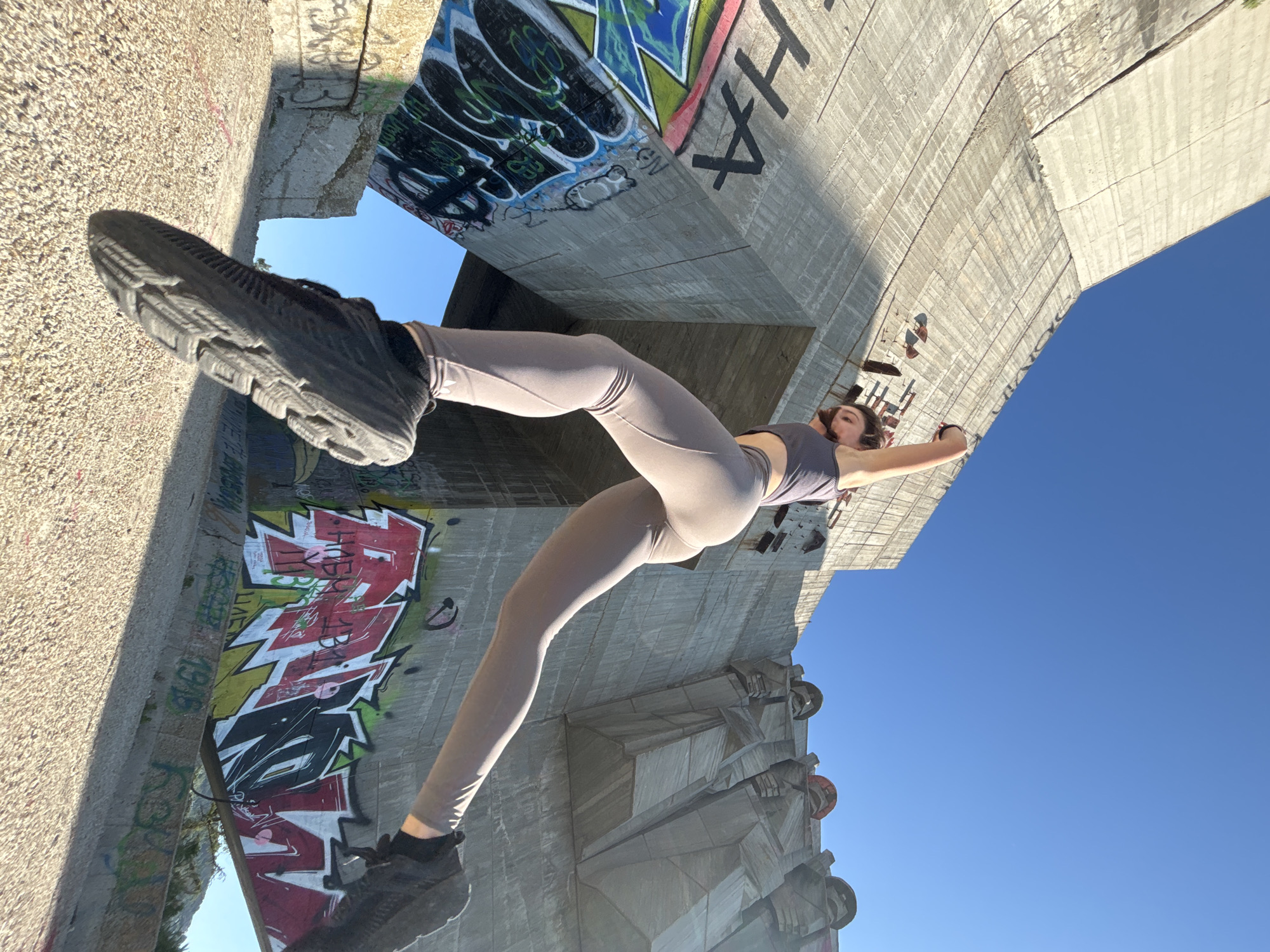 A woman in athletic wear is captured mid-leap in front of the Monument to the Bulgarian-Soviet Friendship in Varna, Bulgaria. The monument, a large brutalist concrete structure, is covered in graffiti. The woman is wearing a dark grey sports bra and light grey leggings. Her right arm is raised above her head, and her left arm is bent at the elbow. Her legs are extended as she jumps. The sky is clear and blue. The ground beneath her is rocky and uneven. The image is taken from a low angle, emphasizing the scale of the monument and the dynamism of the woman's pose.