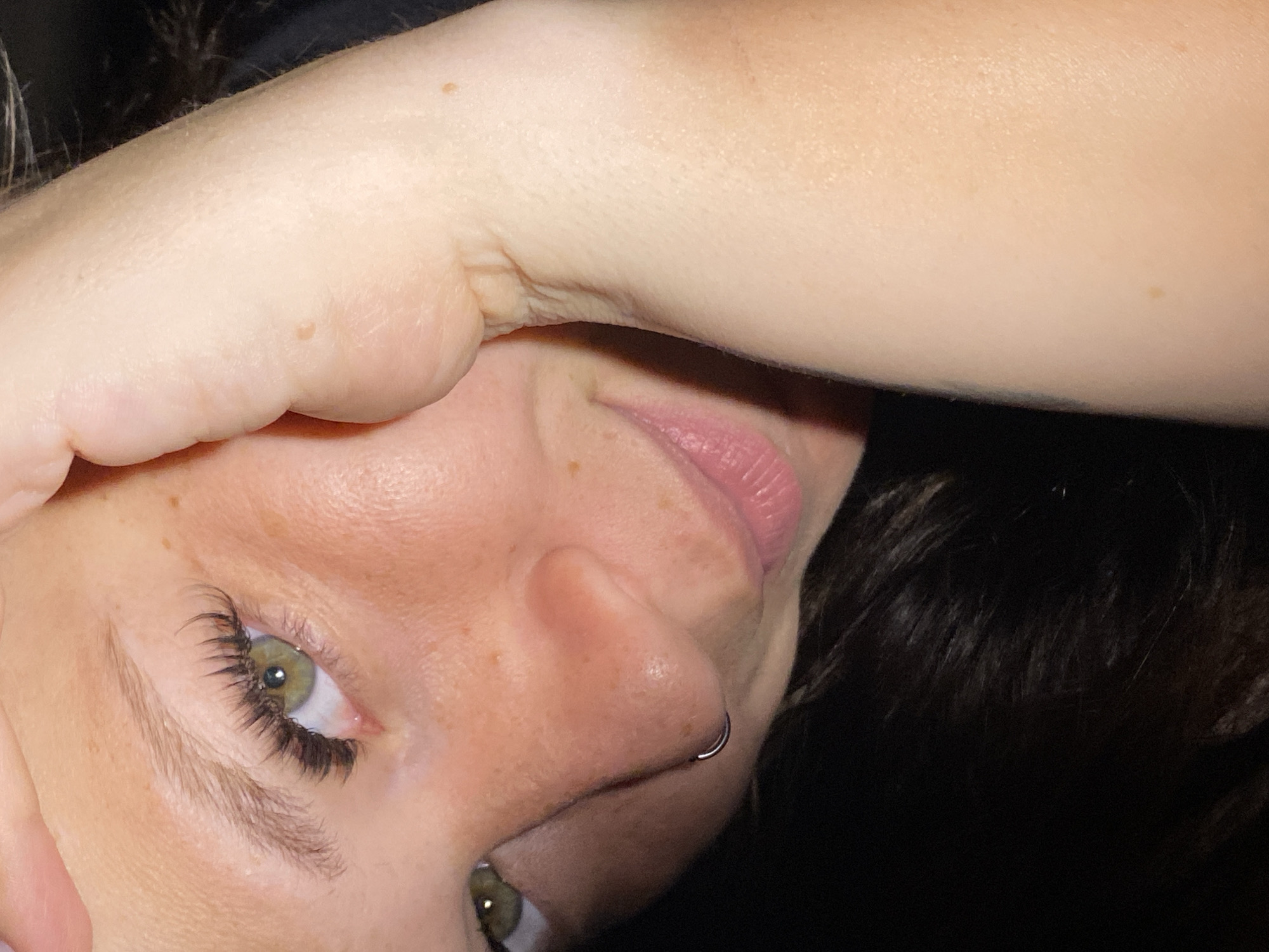The image is a close-up, low-angle shot of a young woman's face. She has green eyes, light skin with freckles, and dark hair. Her right hand is raised, obscuring part of her face and forehead. She is wearing false eyelashes and a small silver hoop piercing in her left nostril. The background is dark, suggesting an indoor setting or low lighting conditions. The image does not contain any information about the location in the city of Chaikovsky, Russia.