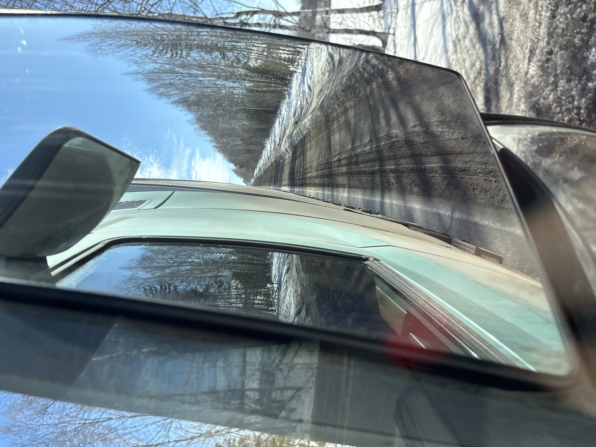 The image presents a roadside view from a moving vehicle in Kukui, Russia. Visible in the left frame is the light teal side panel of a large vehicle, alongside an interior window frame. The prominent right-hand side mirror, soiled with road grime, reflects a receding two-lane road. This road surface is dark, indicating melting snow and dirt, with distinct tire tracks. Flanking the road are snow-covered shoulders transitioning to a dense stand of mostly bare deciduous trees, including apparent birches, with some evergreen specimens interspersed. The sky is bright blue with scattered cirrus clouds, indicative of a clear, sunny day in a late winter or early spring environment. Direct sunlight casts visible shadows. No individuals are depicted.