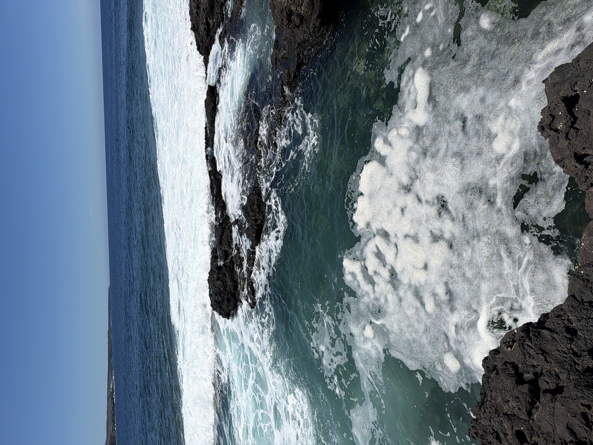The image shows a rocky coastline near Tinajo, Spain. Dark, jagged rocks line the edge of the ocean, where waves crash and create white foam. The water is a mix of deep blue and turquoise. A clear blue sky is visible above the ocean. The angle of the shot is from above, looking down onto the water and rocks.