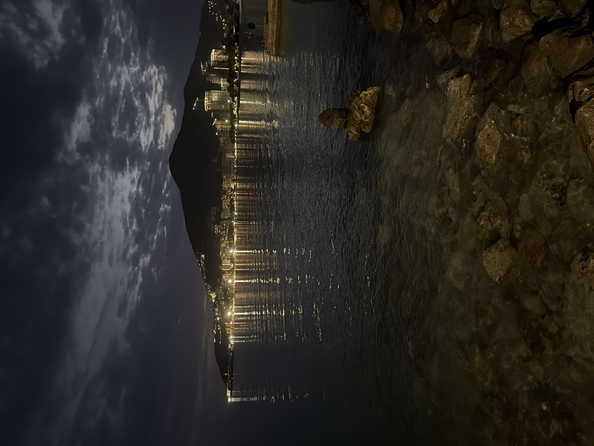 The image presents a captivating nighttime scene of a coastal area in Budva, Montenegro. The sky, occupying the upper left portion, is a dark expanse dotted with dramatic, textured clouds, some appearing lighter due to ambient light. Below the sky, a dark, imposing mountain or hill forms a silhouette against the horizon, anchoring the left and central background.
Along the base of this landform, numerous multi-story buildings, likely part of a town or city, line the coast. These structures are brightly illuminated, with countless windows casting a warm, golden glow. These city lights create prominent, elongated reflections on the calm, dark surface of the water that dominates the mid-ground. The reflections appear as shimmering, vertical streaks, extending from the shoreline across the water.
In the immediate foreground on the right, a rugged, rocky shoreline is visible. Large, dark rocks, some fully exposed and others partially submerged in the clear, shallow water, add a natural, textured element to the scene. The water around these rocks is still and appears transparent.
The time of day is clearly night, emphasized by the dark sky and the reliance on artificial lighting. The weather appears calm, with the clouds adding a sense of depth to the night sky without suggesting adverse conditions. No people or visible text are present in the image. The scene evokes a serene and picturesque atmosphere, highlighting the beauty of urban lights reflected on tranquil water against a natural backdrop.