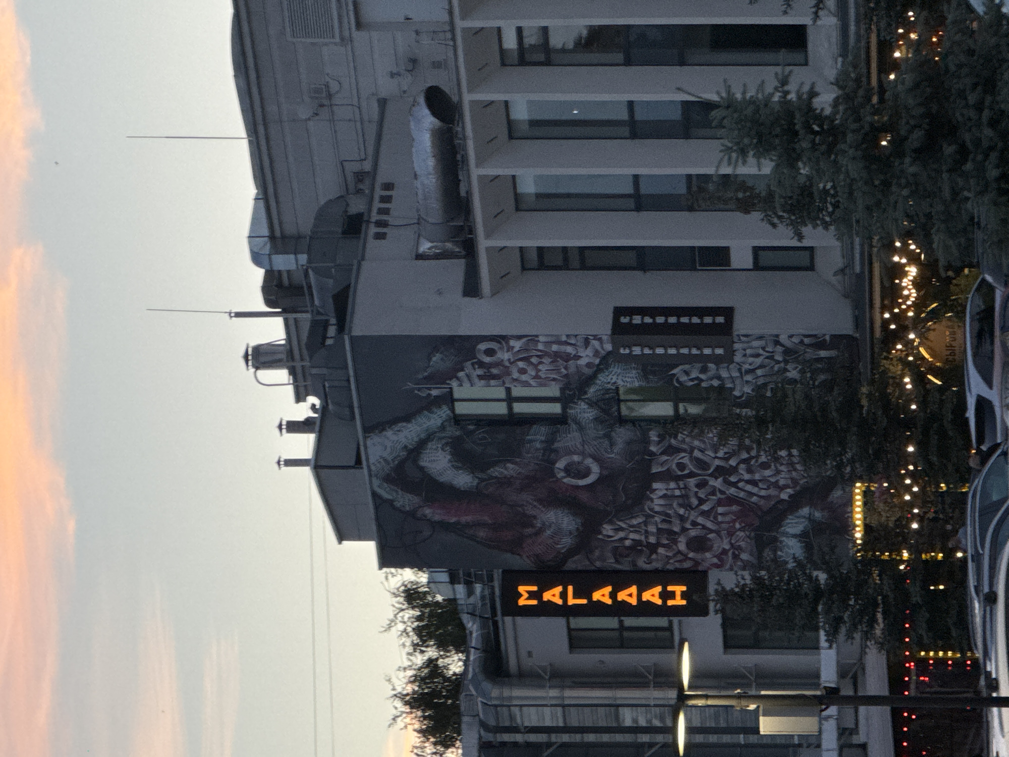 The image shows a building in Nizhny Novgorod, Russia. The building has a large mural on its side. There are also signs on the building, one of which reads "МАЛАААН" in orange neon letters. Other signs on the building are written in Cyrillic script. In the foreground, there are trees and cars. The sky is a pale pink and orange, suggesting it is either sunrise or sunset. The architecture of the building appears to be modern, with large windows and metal ventilation systems visible.