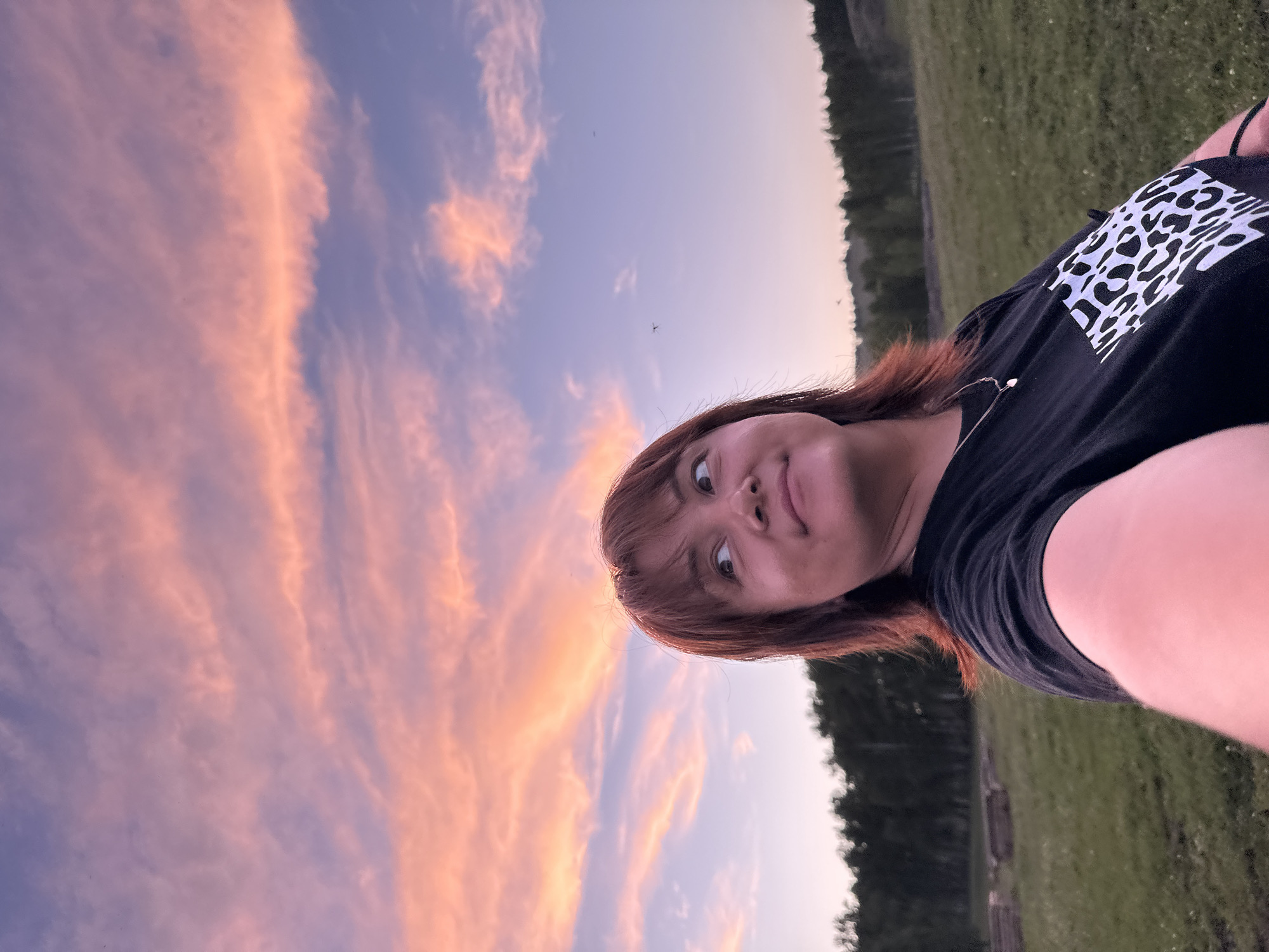 A woman with shoulder-length auburn hair looks up and to the right, her face angled upwards and away from the camera. She is wearing a black t-shirt with a white and black leopard print design on the left side of her chest and a silver necklace with a small pendant. The background shows a grassy field with a line of trees in the distance and a sky filled with pink and purple clouds, suggesting it is either sunrise or sunset. The location is identified as Unknown, Unknown.