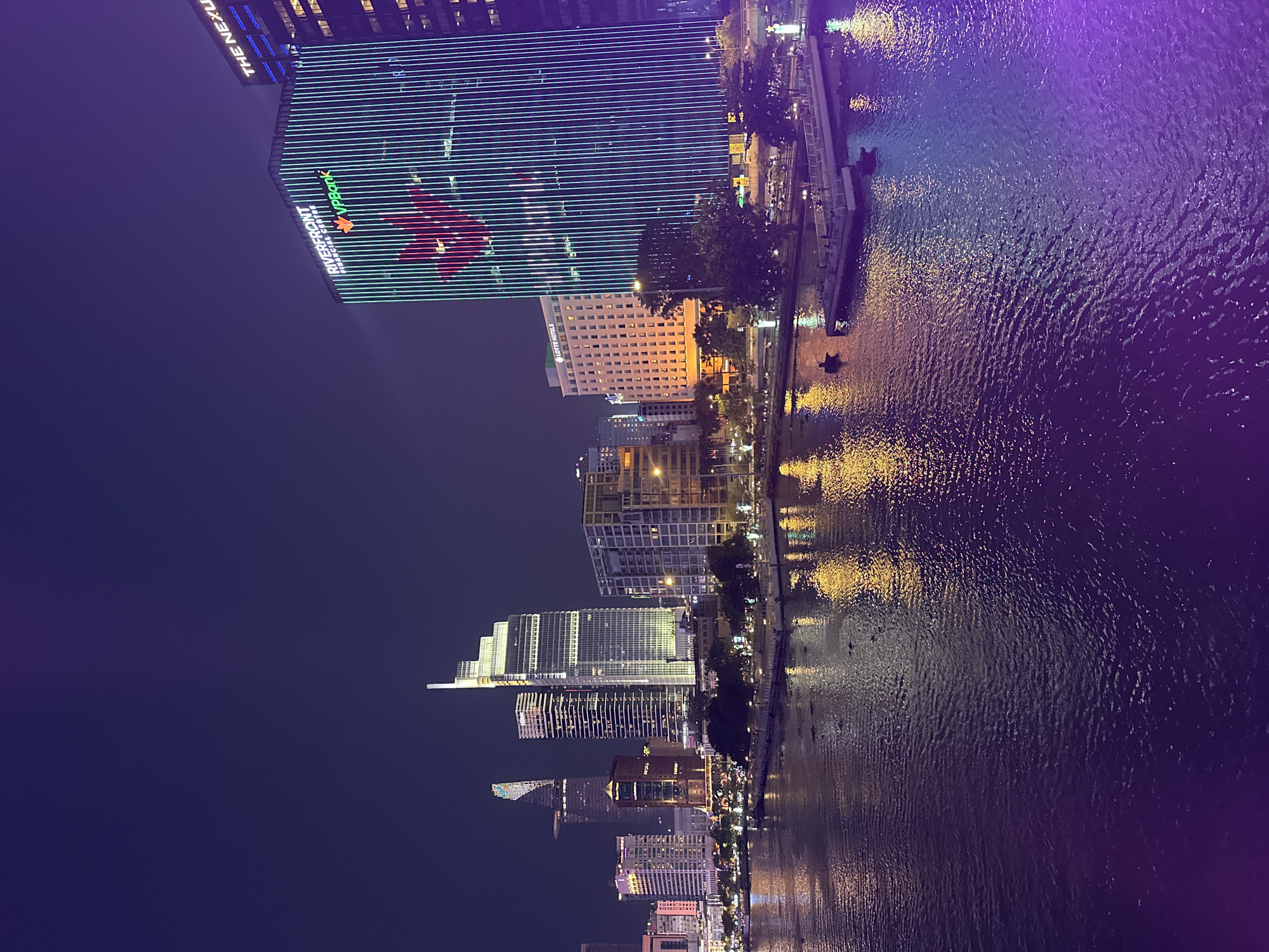 This is a nighttime shot of the Thủ Đức city skyline in Vietnam. The Saigon River flows in the foreground, reflecting city lights. High-rise buildings dominate the skyline, including the Landmark 81 building with a red star logo and linear blue-green lighting. Other buildings are also illuminated, creating a vibrant urban landscape. The Saigon River waterfront is visible along the riverbank.