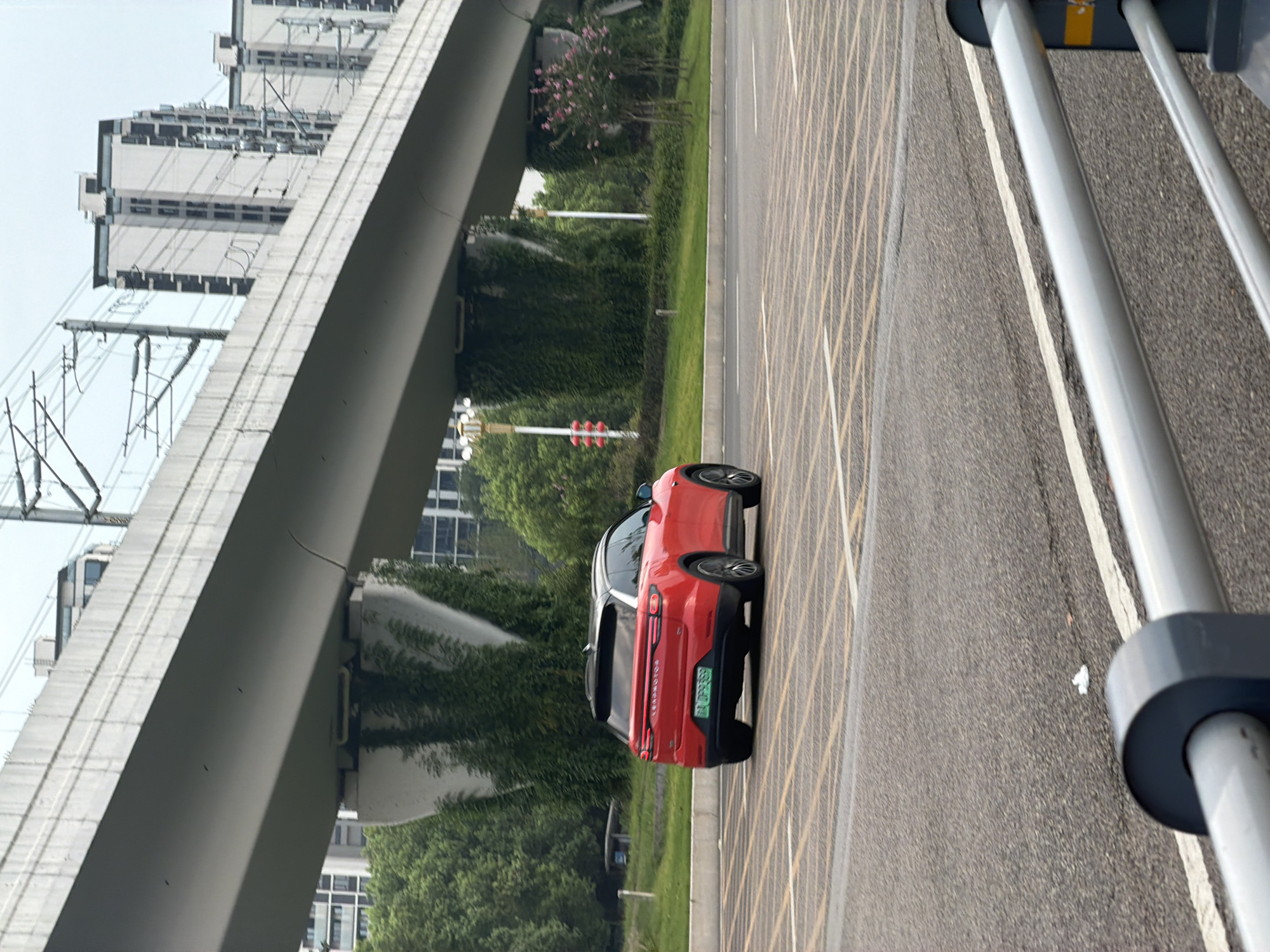 A red electric vehicle, a Leapmotor C11, is captured from the rear-left perspective in motion on a paved road. The car's license plate is visible and reads "JGD359". The road surface is marked with yellow diagonal lines, indicative of a parking area or a special lane. In the background, a massive elevated highway structure dominates the scene, with its concrete pillars covered in lush green ivy. Beyond the highway, modern high-rise apartment buildings are visible, some with dark window frames. Trees and grassy areas are present along the roadside. A traffic light, showing red, is situated to the left of the car. The sky is overcast. The image was taken in the city of Jiangjin, China.