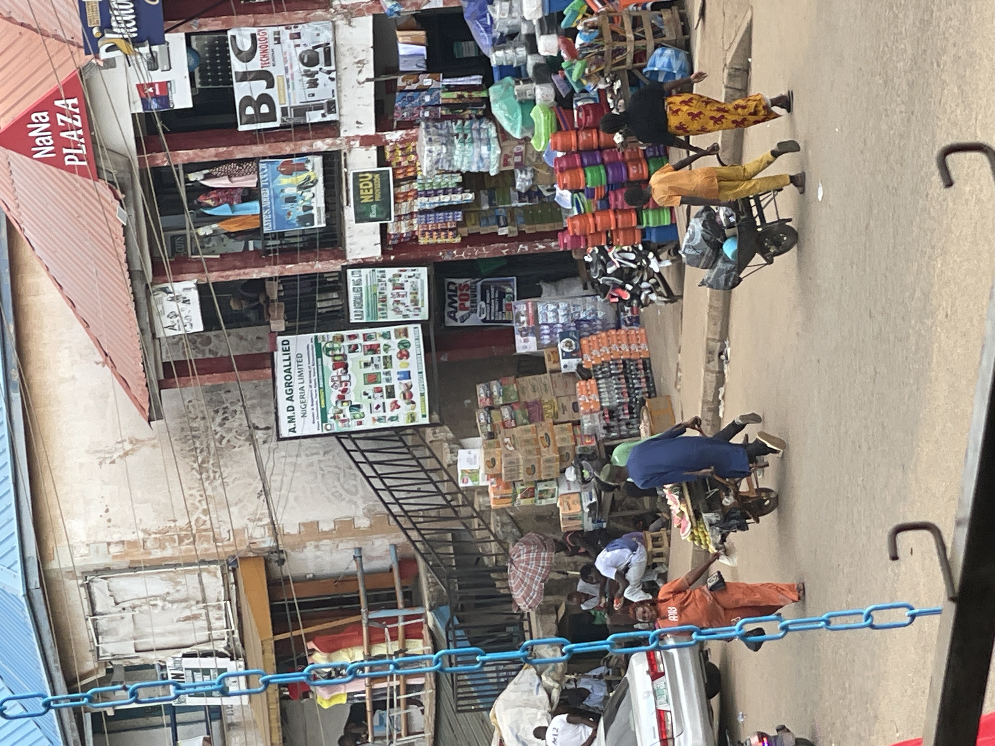 The image depicts a bustling street scene in an outdoor marketplace. A bright blue chain hangs prominently in the foreground, suggesting some form of scaffolding or temporary structure. The background features a multi-story building with various shops and signage. One sign reads "NaNa PLAZA." The building's facade is weathered, with peeling paint and exposed brickwork in places. A staircase with black metal railings leads up to an upper level.
In the middle ground, people are engaged in various activities. A woman in an orange jumpsuit pushes a cart laden with produce, possibly fruits. To her right, a person in a blue robe pushes a similar cart filled with bags. Another person in an orange shirt and yellow pants is seen pulling a cart carrying black bags. Several other individuals are visible in the background, some standing and others seated. Stalls are set up with an array of goods, including stacks of boxes, plastic containers of various colors, and bottles of beverages. A red and white checkered umbrella is open, providing shade. A silver car is parked on the left side of the street.
The overall environment appears to be a busy commercial area, with a mix of goods and people interacting within a somewhat worn urban setting. The lighting suggests it is daytime.