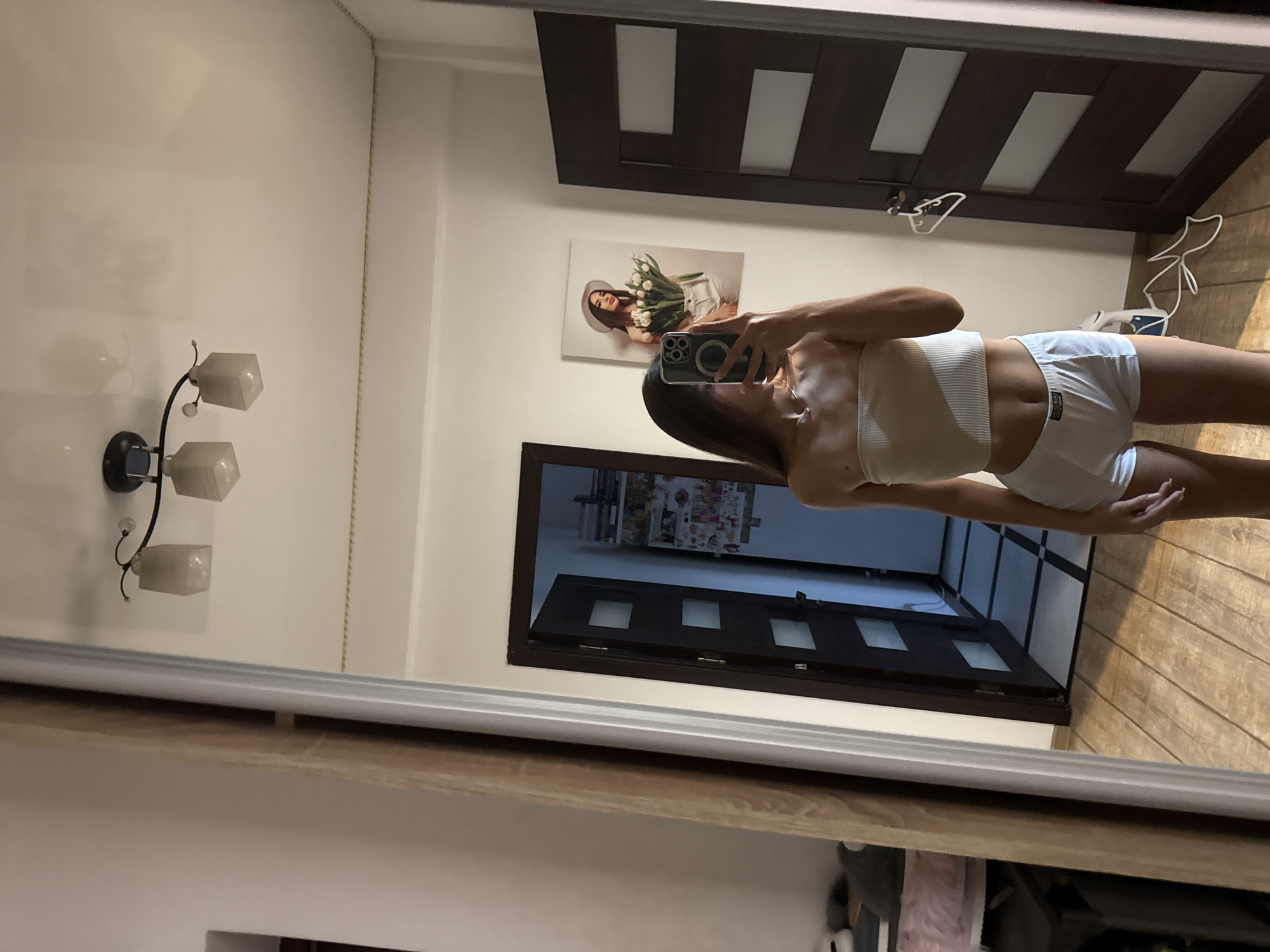 A young woman with long brown hair is taking a mirror selfie in a room. She is wearing a beige bandeau top and white shorts. The room appears to be a bedroom or dressing area, with a large mirror, two dark wood doors, and wooden flooring. There is a painting of a woman holding flowers on the wall behind her. The woman is holding a smartphone in her right hand, and a magnetic phone accessory is visible on the back of the phone. A ceiling light fixture with two square glass shades is visible at the top of the mirror's reflection. A clothing rack with a hanger is attached to one of the doors. The overall impression is of a private, domestic space.