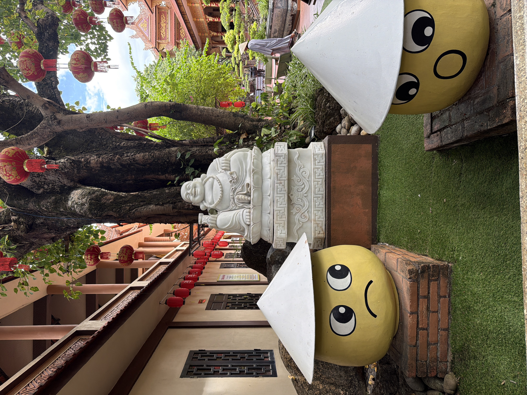 The image depicts a courtyard within a Buddhist temple complex in Hoi An, Vietnam, likely the Ong Pagoda (Chua Ong) due to the architectural style and decorative elements. In the foreground, two large, yellow, decorative figures resembling plump fruit are positioned. The figure on the left has a sad facial expression with downturned eyes and a downturned mouth, wearing a conical straw hat. The figure on the right has a surprised expression with wide, circular eyes and an open mouth, also wearing a conical straw hat. Behind these figures, a white stone statue of a laughing Buddha is centrally placed on a tiered pedestal. A large, gnarled tree with lush green foliage rises behind the Buddha statue, adorned with numerous red, cylindrical lanterns, some with gold Chinese characters. The background reveals the ornate architecture of the temple, with red-tiled roofs, intricate carvings, and decorative eaves. A person dressed in a gray robe and a conical hat is seen walking away in the mid-ground, along a pathway lined with manicured bonsai trees and potted plants. The ground is covered with green artificial turf in the immediate foreground and brick paving further back. The overall scene suggests a serene and culturally rich temple environment.