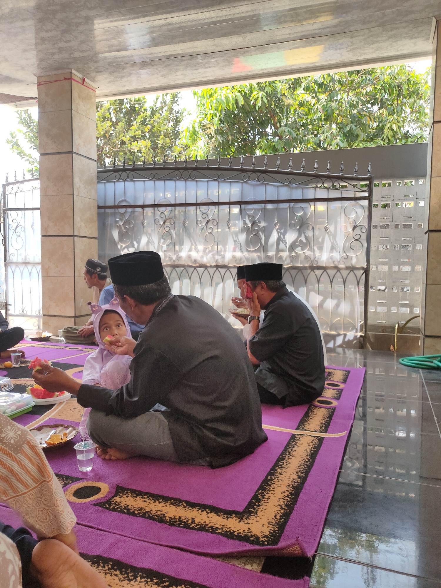 The image shows a gathering of people, likely a family, in an outdoor, covered area in Sumedang, Indonesia. The individuals are seated on a large purple rug with an orange and black border. One man in a black peci (cap) and black shirt is feeding a piece of watermelon to a young child wearing a light purple head covering and a matching garment. The child looks at the man with anticipation. Another man, also wearing a black peci and black shirt, is seated further back, facing away from the camera and appears to be eating. In the background, there is a decorative metal fence and greenery from trees. The floor appears to be tiled. A green hose is visible on the right side of the image. The overall atmosphere seems to be one of a casual, communal meal.